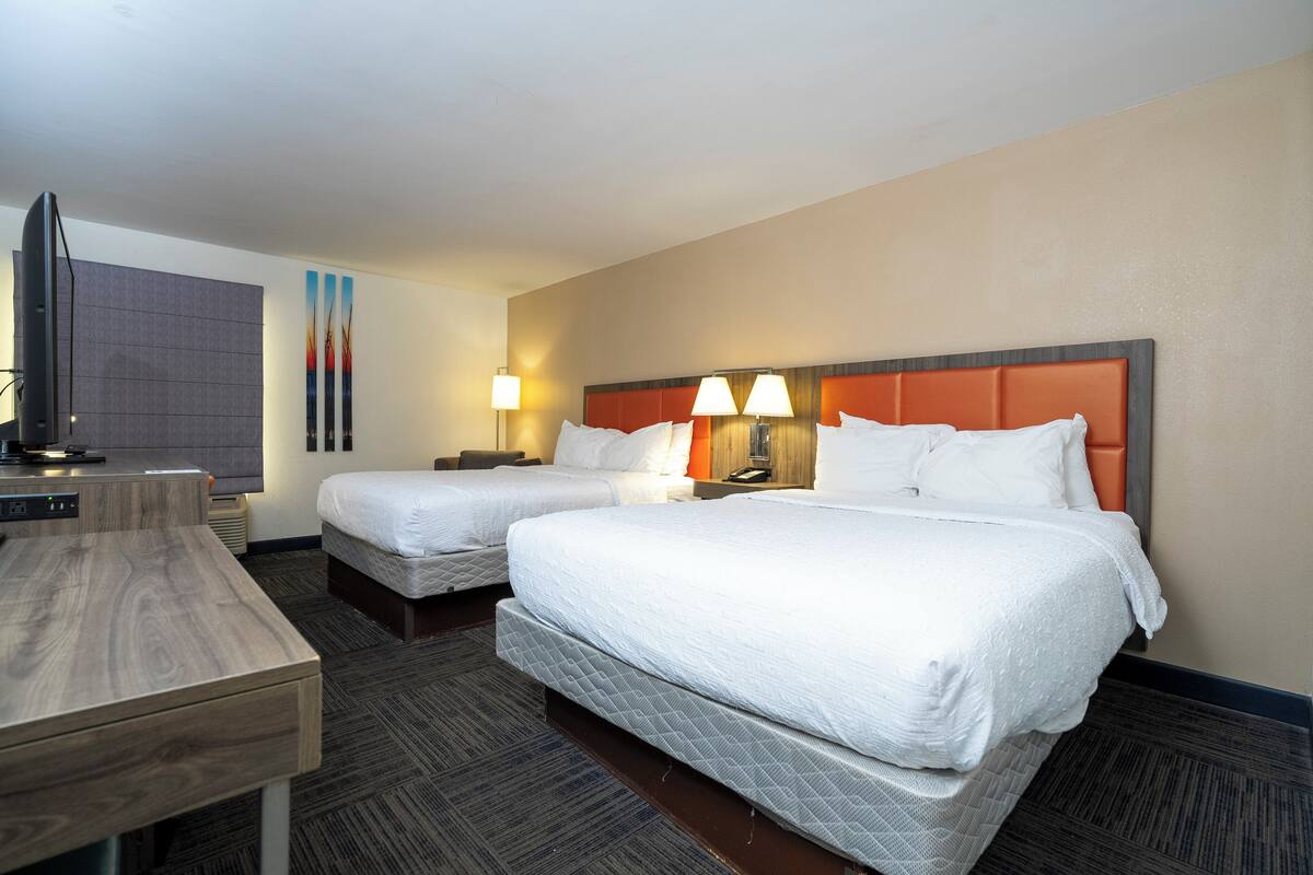 A hotel room with two beds, a desk, a flat-screen TV, and a wall-mounted TV.