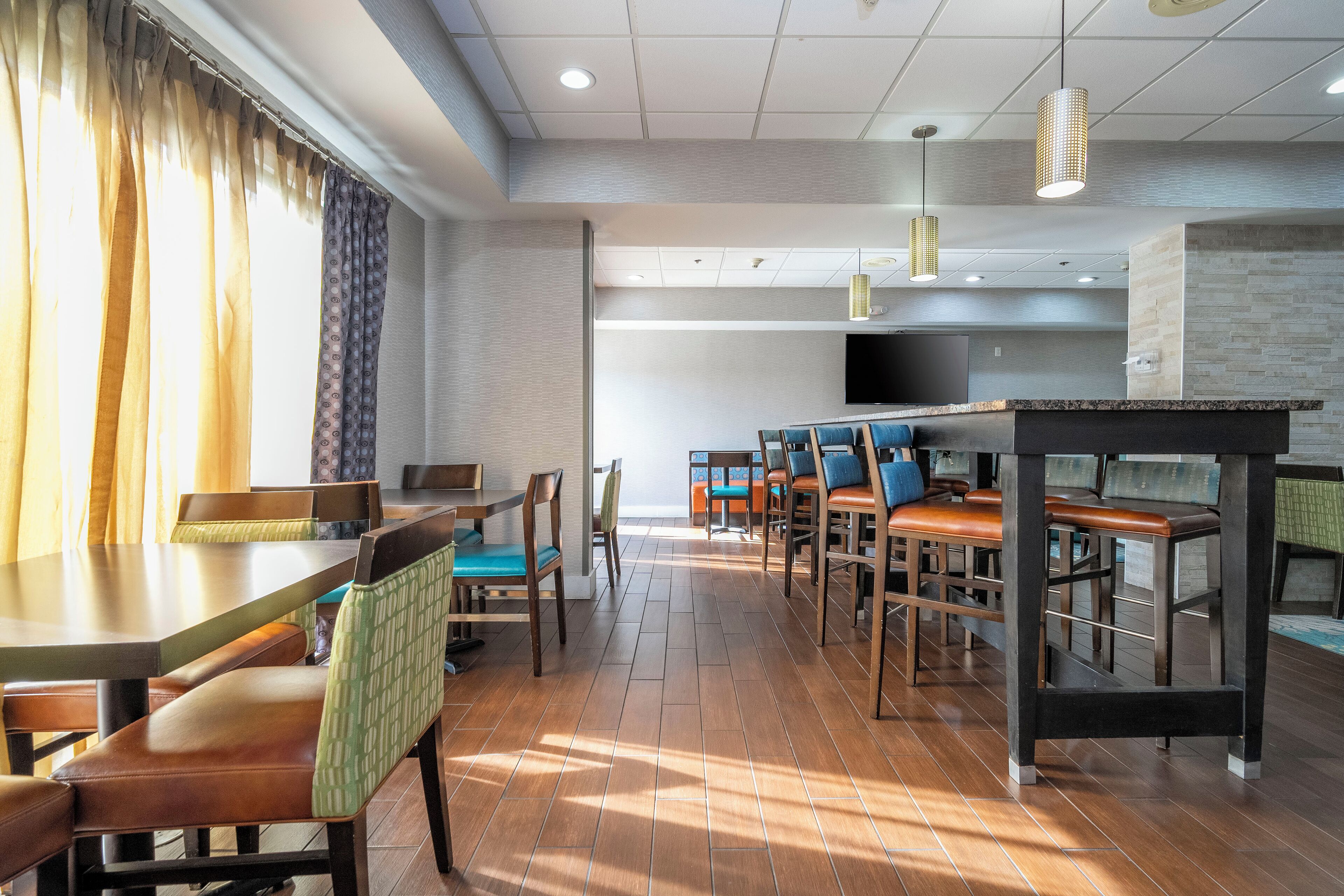 A modern dining area with multiple tables and chairs, a flat-screen TV mounted on the wall, and a large window with curtains.