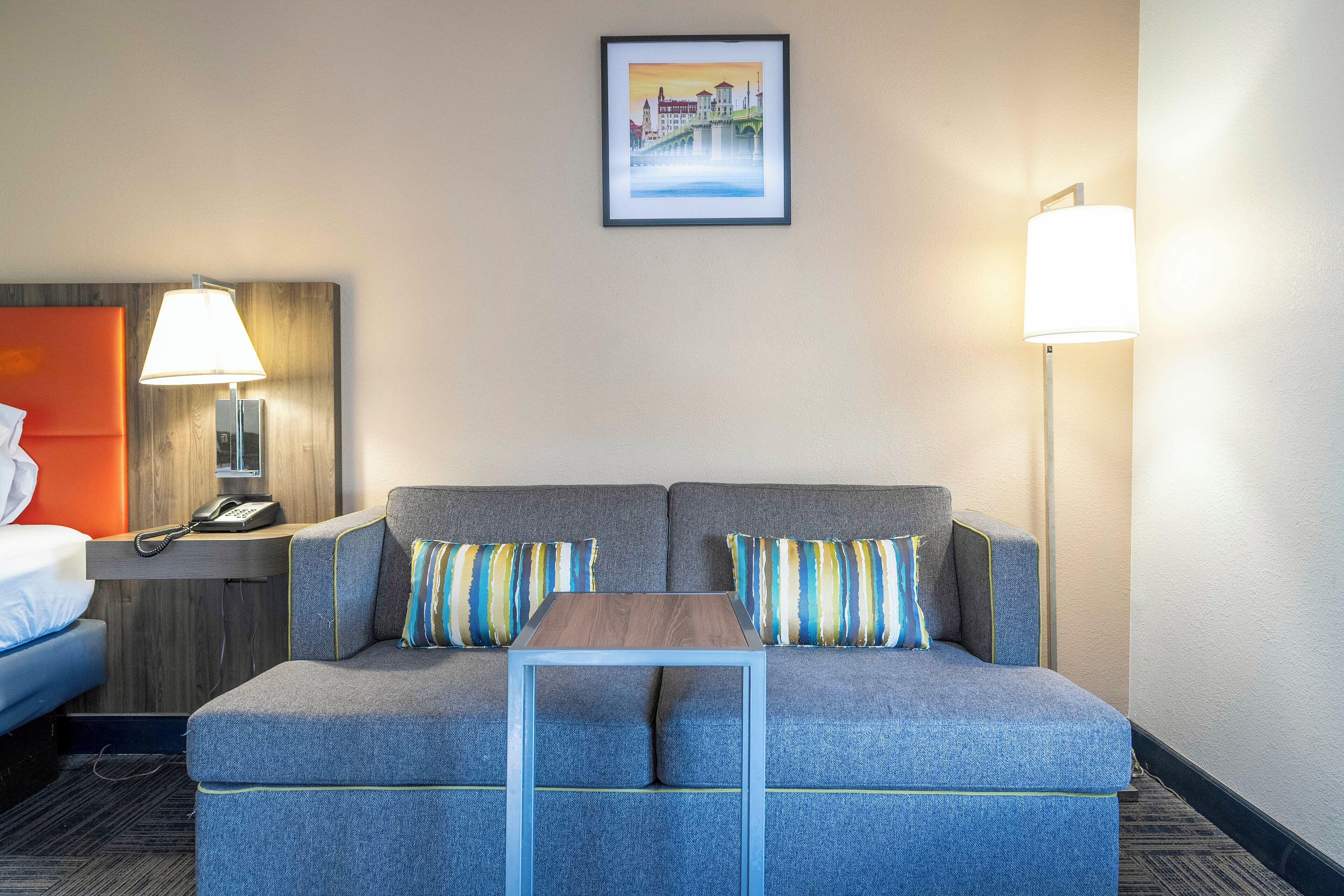 A hotel room with a sofa, a small table, a lamp, a phone, and a picture on the wall.