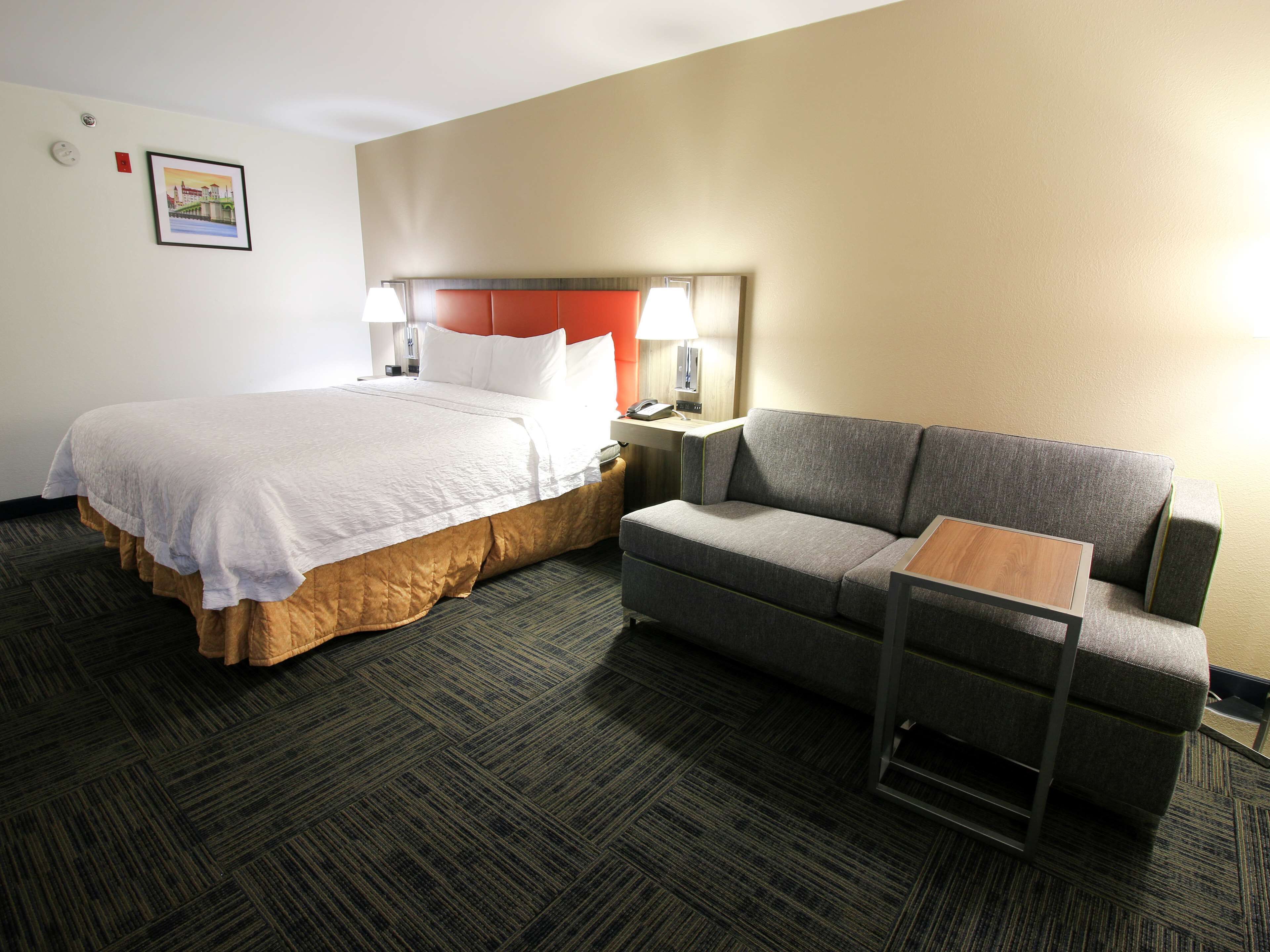 A hotel room with a bed, a gray sofa, a small wooden table, and a nightstand with a lamp.
