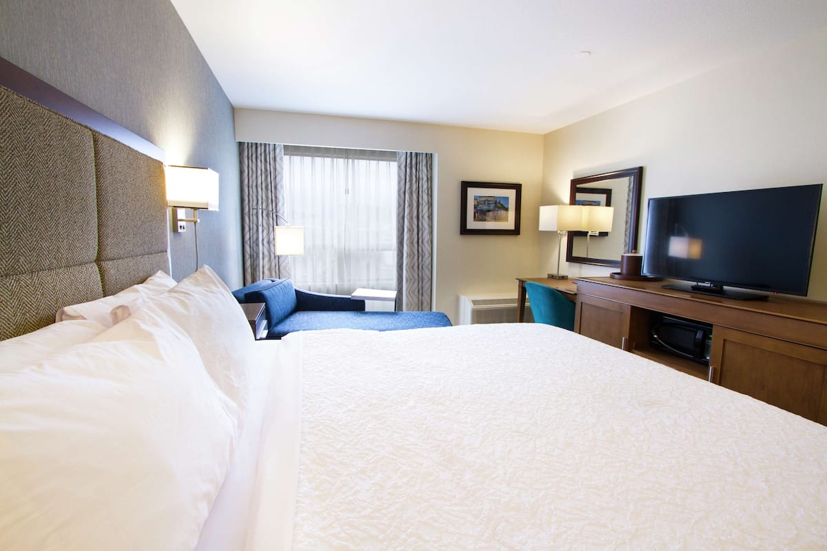 A hotel room with a large bed, a desk with a flat-screen TV, a chair, and a window with curtains.