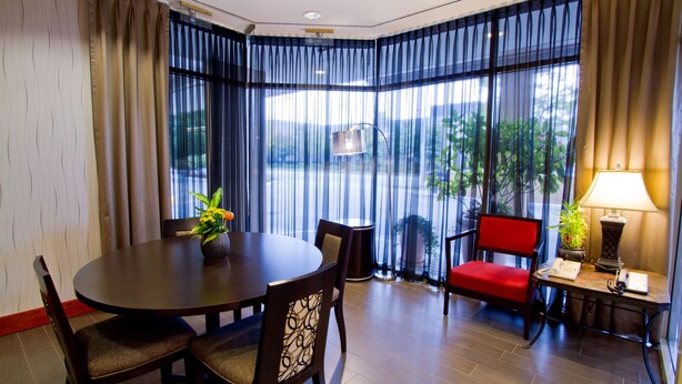 Habitación de hotel con una mesa redonda de comedor, dos sillas, una butaca roja, una mesita con una lámpara y vistas a través de ventanas grandes con cortinas translúcidas.