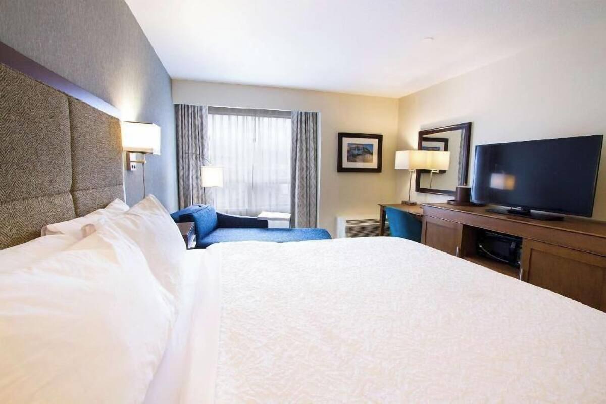 A hotel room with a large bed, a television, a desk with a lamp, and a window with curtains.