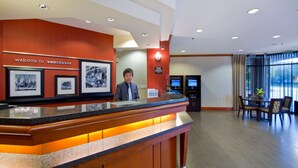 Mostrador de recepción con un cartel que dice "Welcome to Vancouver", cuadros enmarcados, una recepcionista, una pantalla digital y un área de comedor con una mesa y sillas.