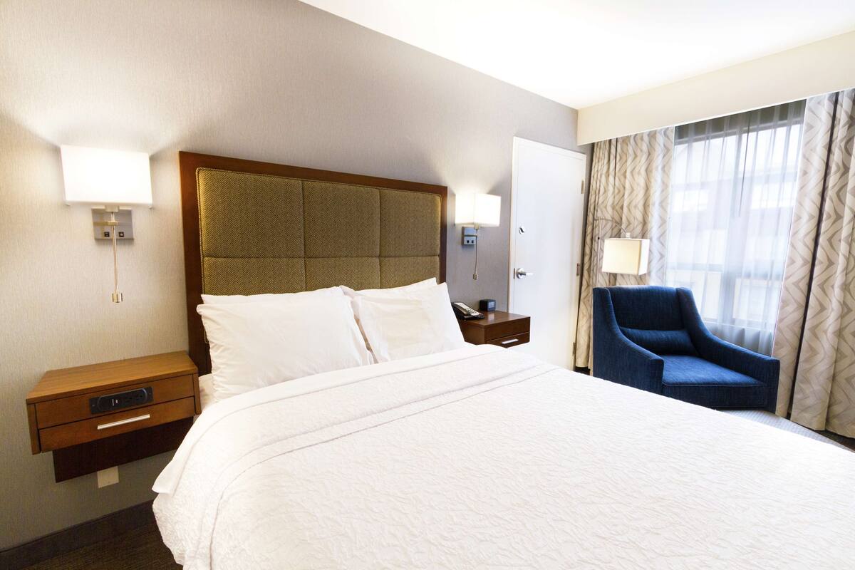 A hotel room with a large bed, a wooden nightstand, a blue armchair, and two wall-mounted lamps.