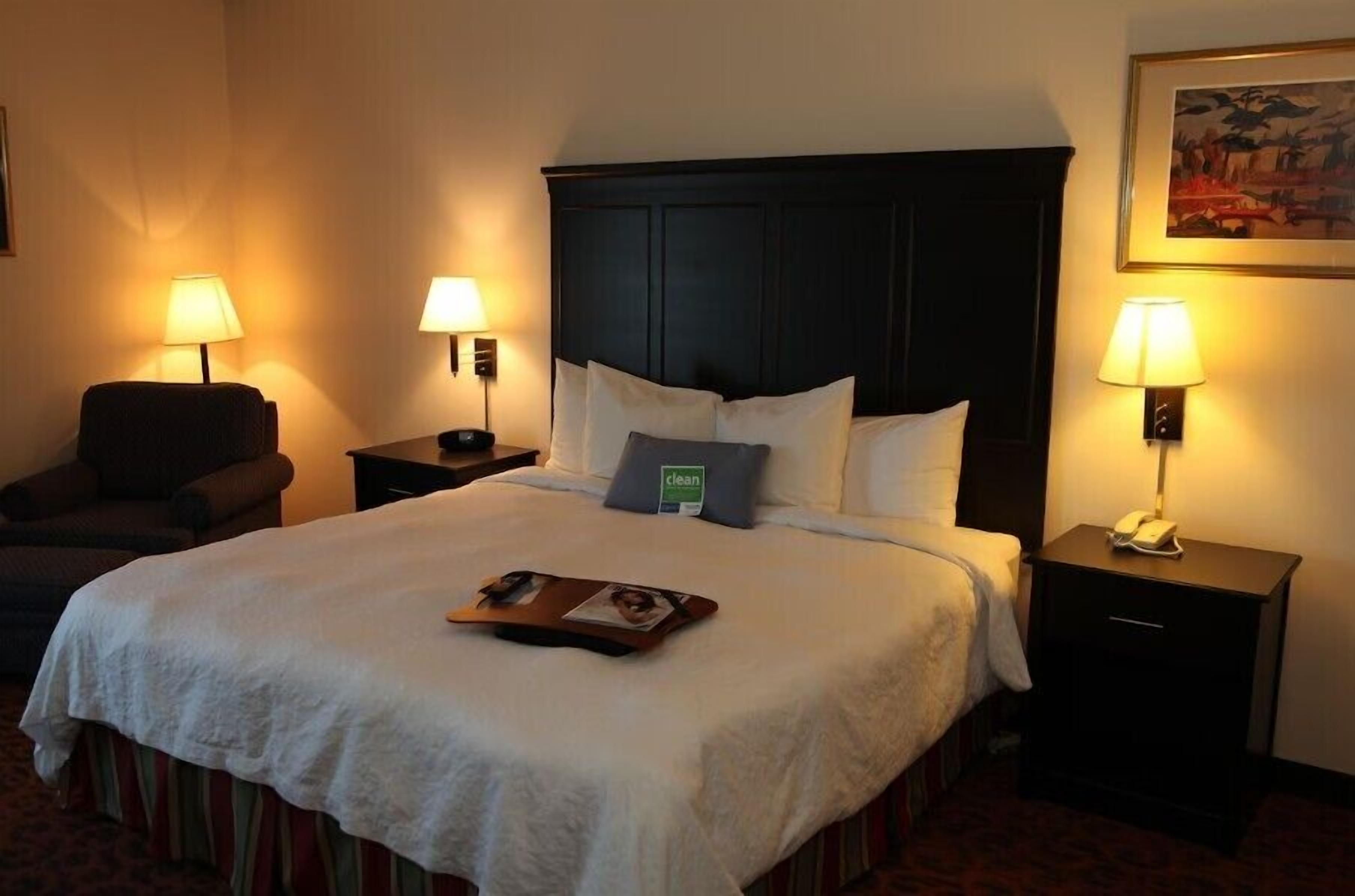 A hotel room with a large bed, two bedside tables with lamps, a chair, and a framed picture on the wall.