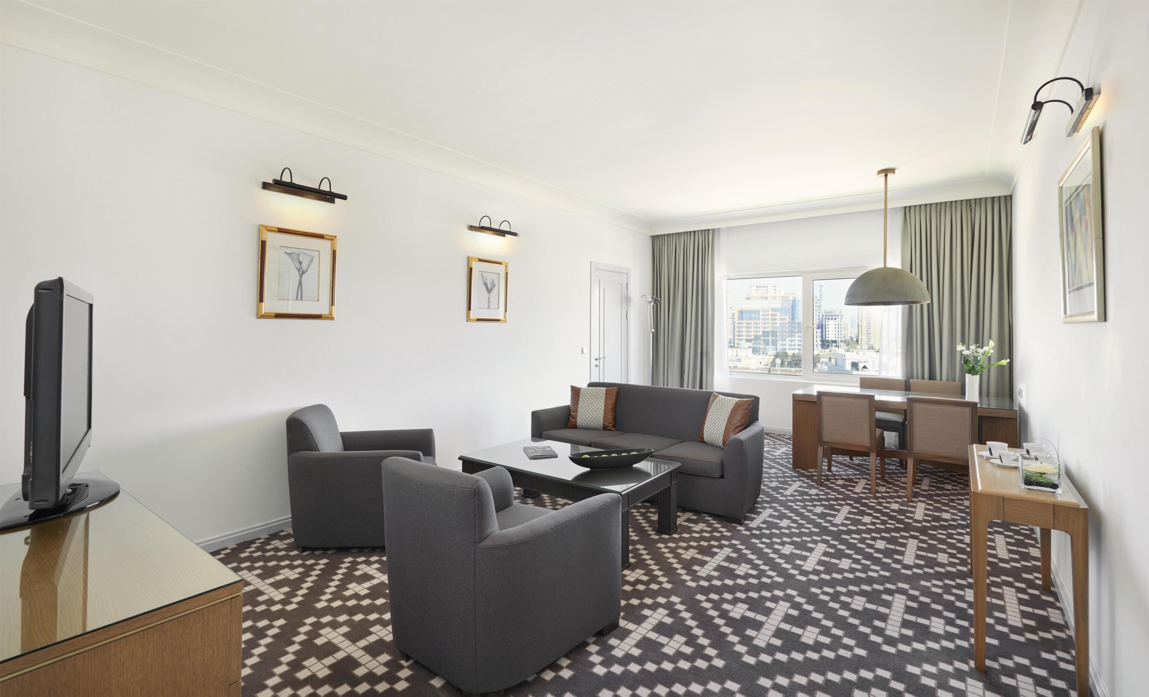 Une chambre d’hôtel moderne comprenant un canapé, des fauteuils, une table basse, un téléviseur, un coin repas et un sol à motifs.