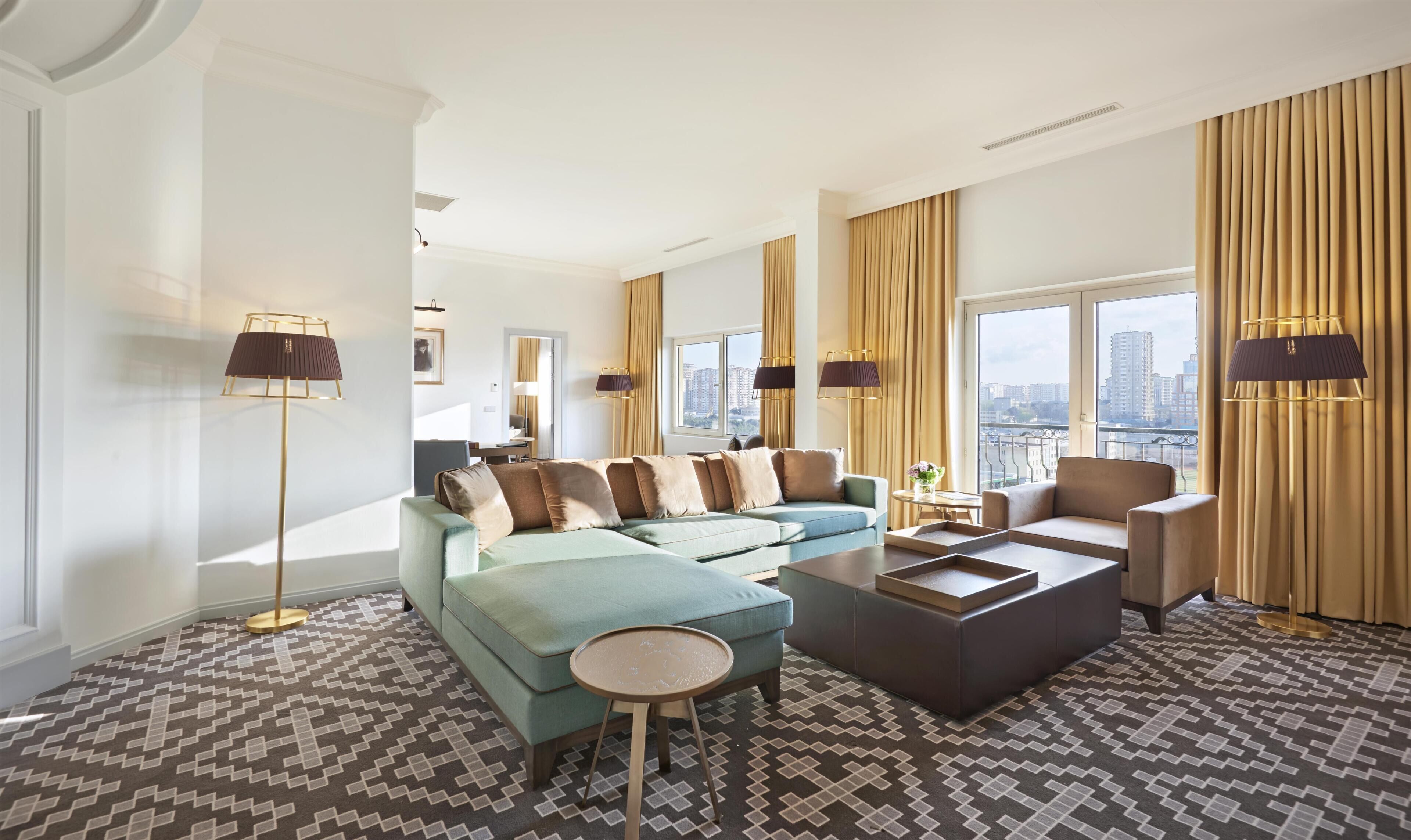 A modern living room with a sectional sofa, a coffee table, and a chair. There are floor lamps and a view of the city through large windows.