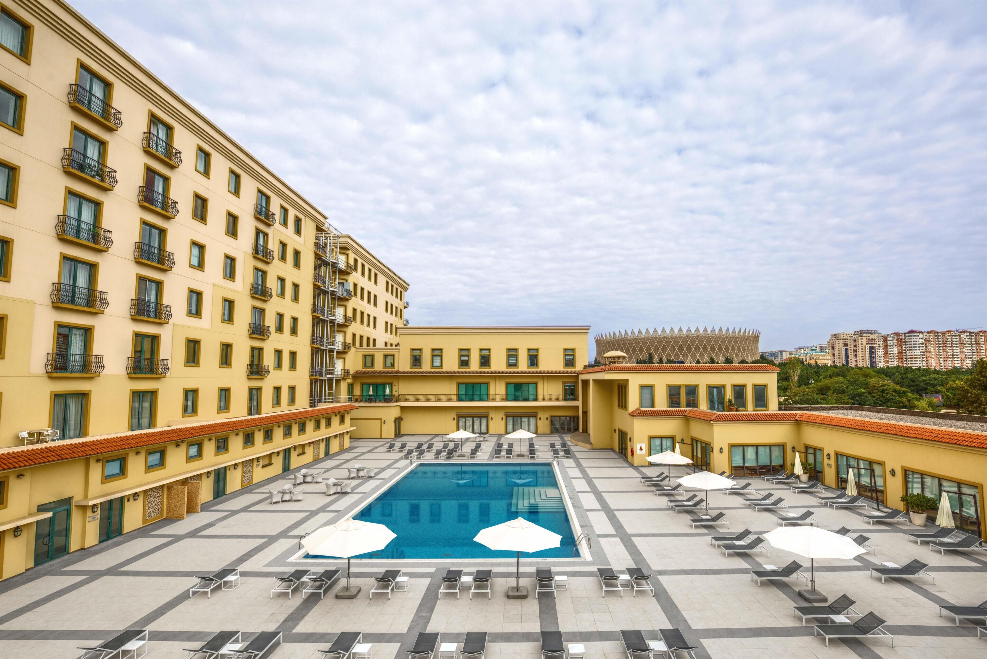 A hotel with a central swimming pool, multiple balconies, and a courtyard with lounge chairs and umbrellas.
