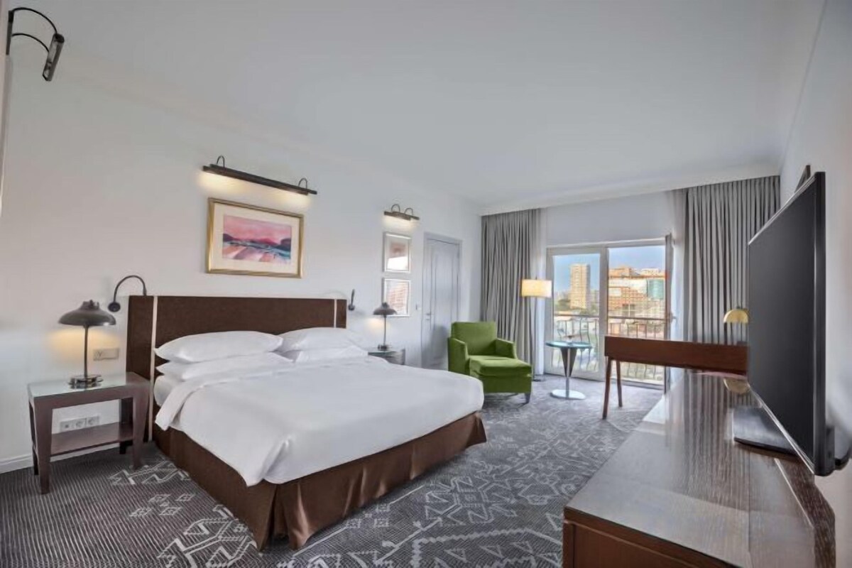 A hotel room with a large bed, a television, a desk, and a view of the city through the window.