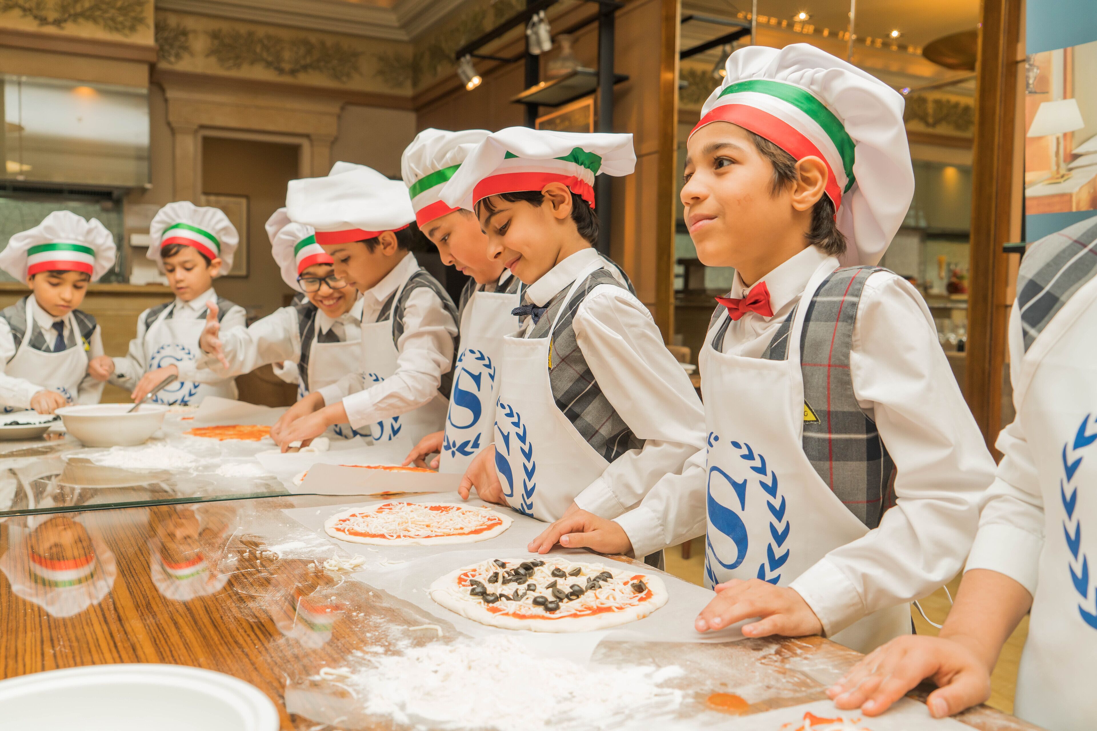 A group of children in chef hats and aprons preparing pizzas.