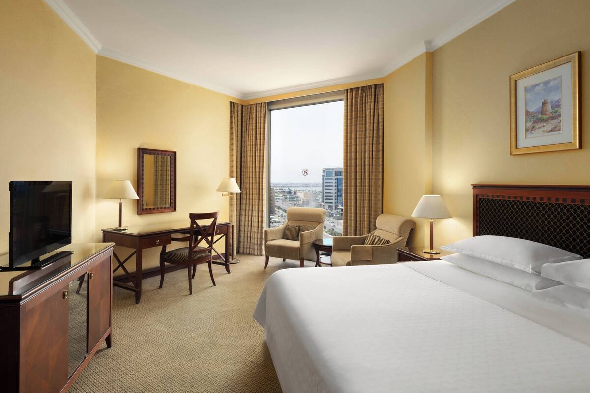 A hotel room with a large bed, a desk with a chair, a television, and a view of the city through the window.