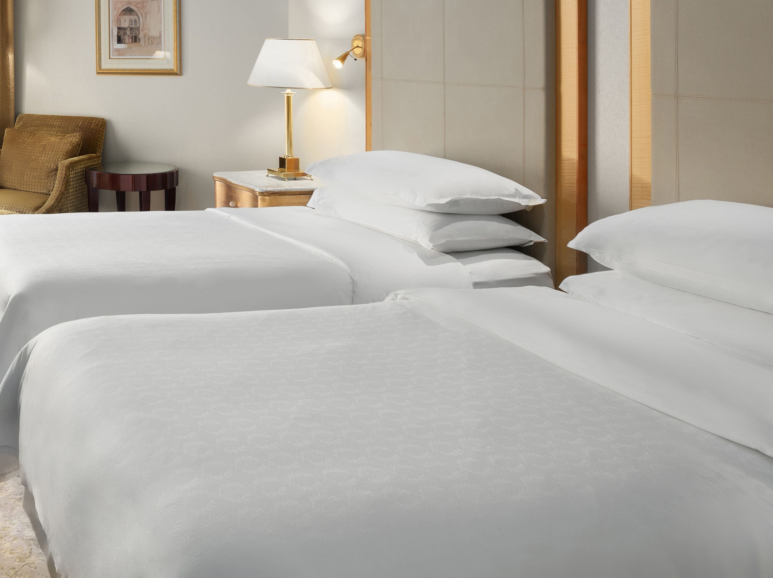 A hotel room with two beds, a bedside table with a lamp, a framed picture on the wall, and a chair.