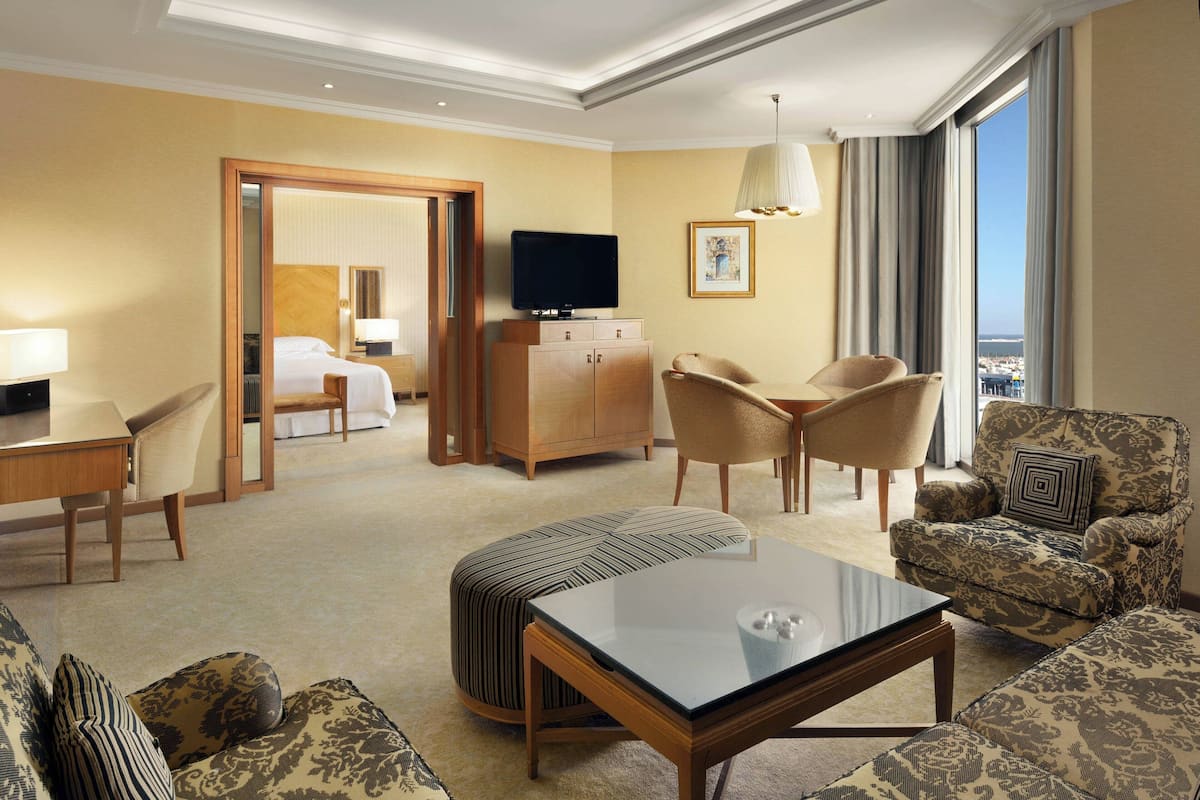 A hotel room with a sofa, armchair, ottoman, a television on a wooden cabinet, and a large window with a view.