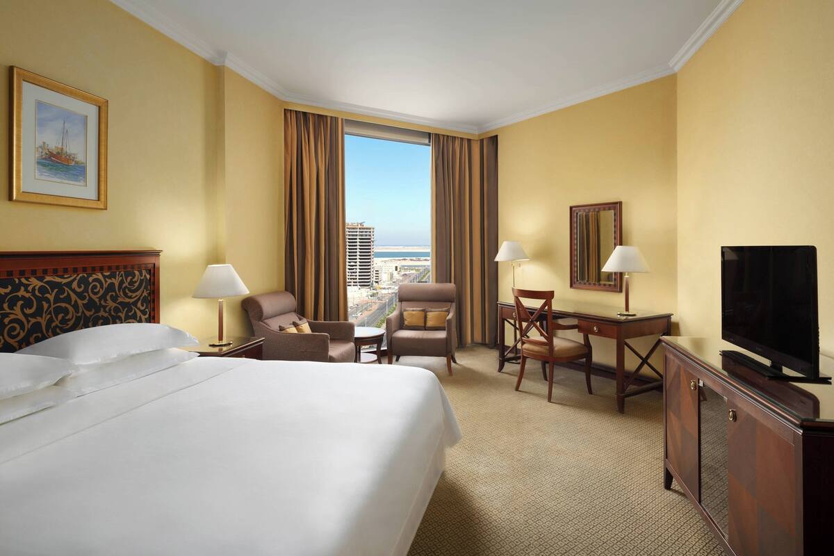 A hotel room with a large bed, a desk, a chair, a television, and a view of the city.