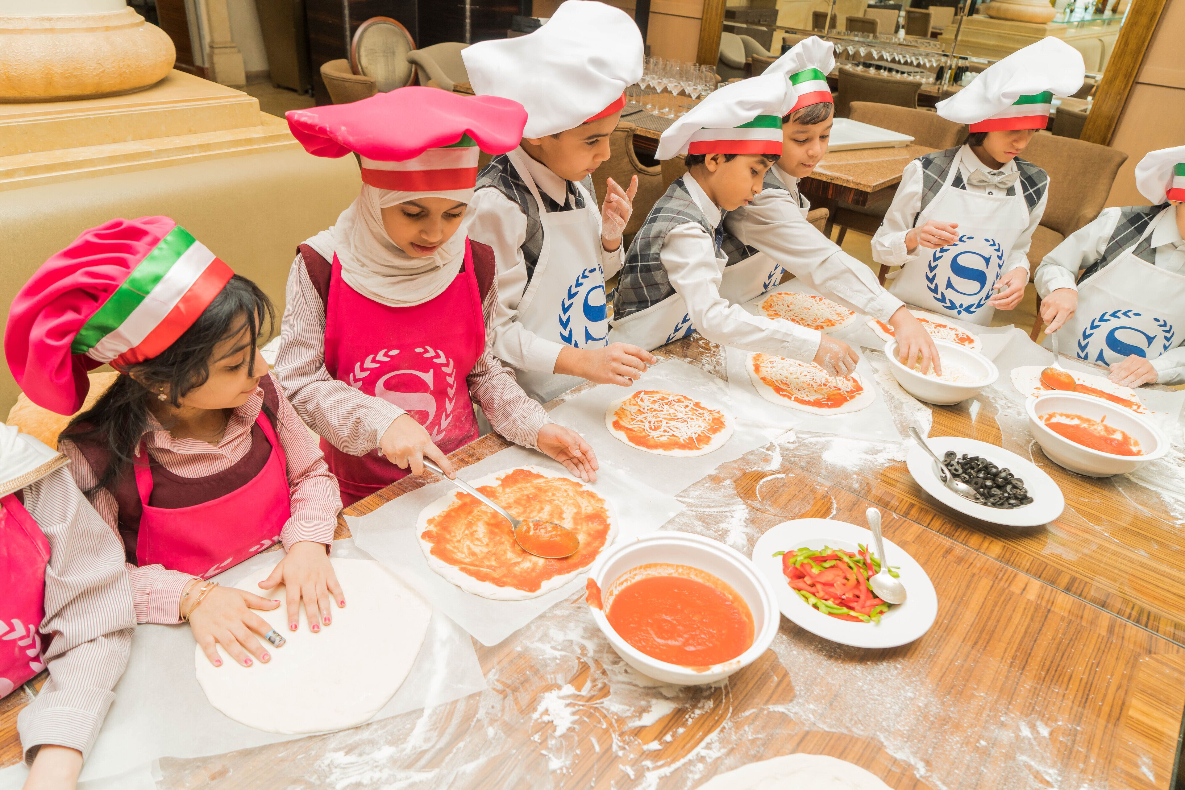 Children in chef hats making pizza in a kitchen.