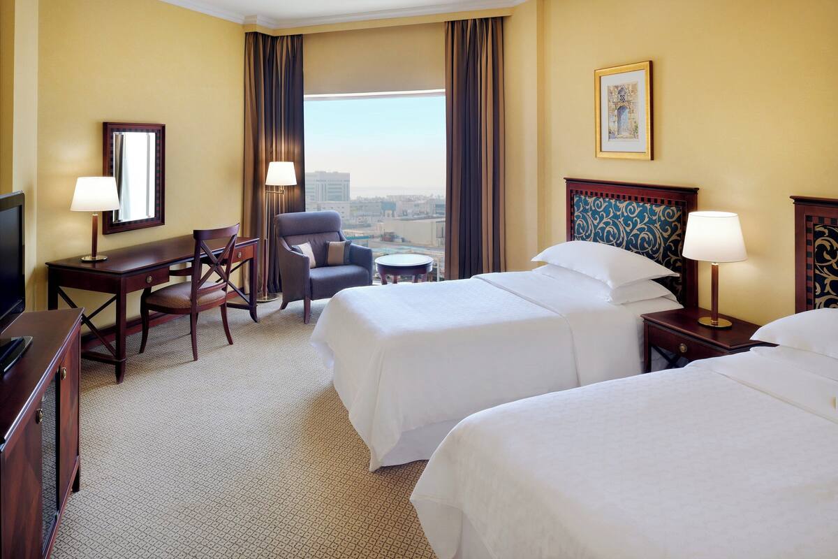A hotel room with two beds, a desk, a chair, a small round table, and a television.