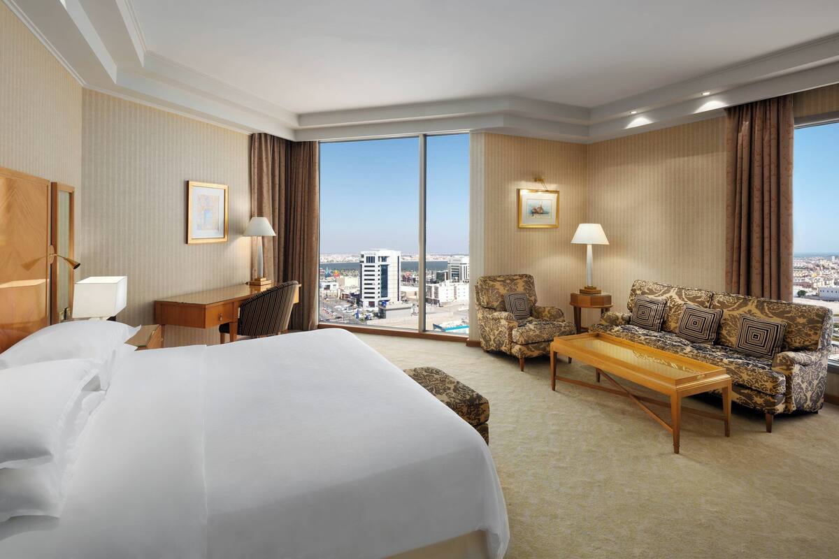 A hotel room with a large bed, a sofa, a coffee table, a desk, and a chair. There are two windows with curtains, and a city view is visible through the windows.