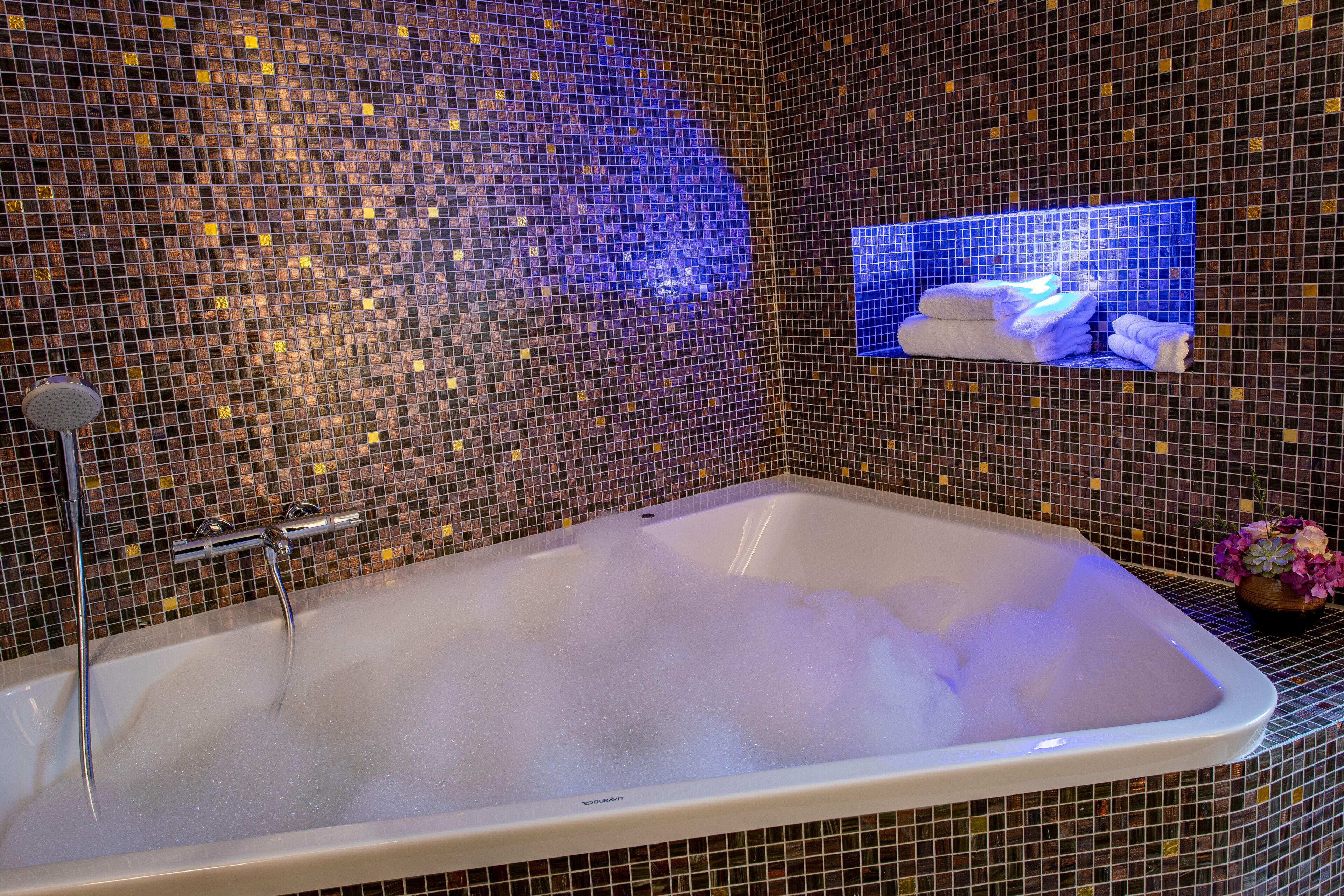 A large bathtub with a mosaic tile wall, a showerhead, and a shelf with towels.