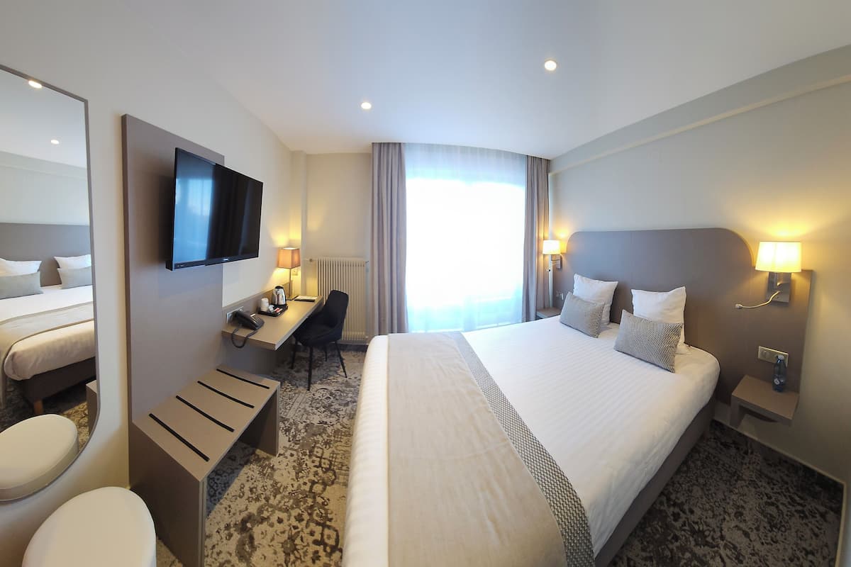 Une chambre d’hôtel avec un grand lit, un bureau, une chaise, des toilettes, une télévision et une fenêtre avec des rideaux.
