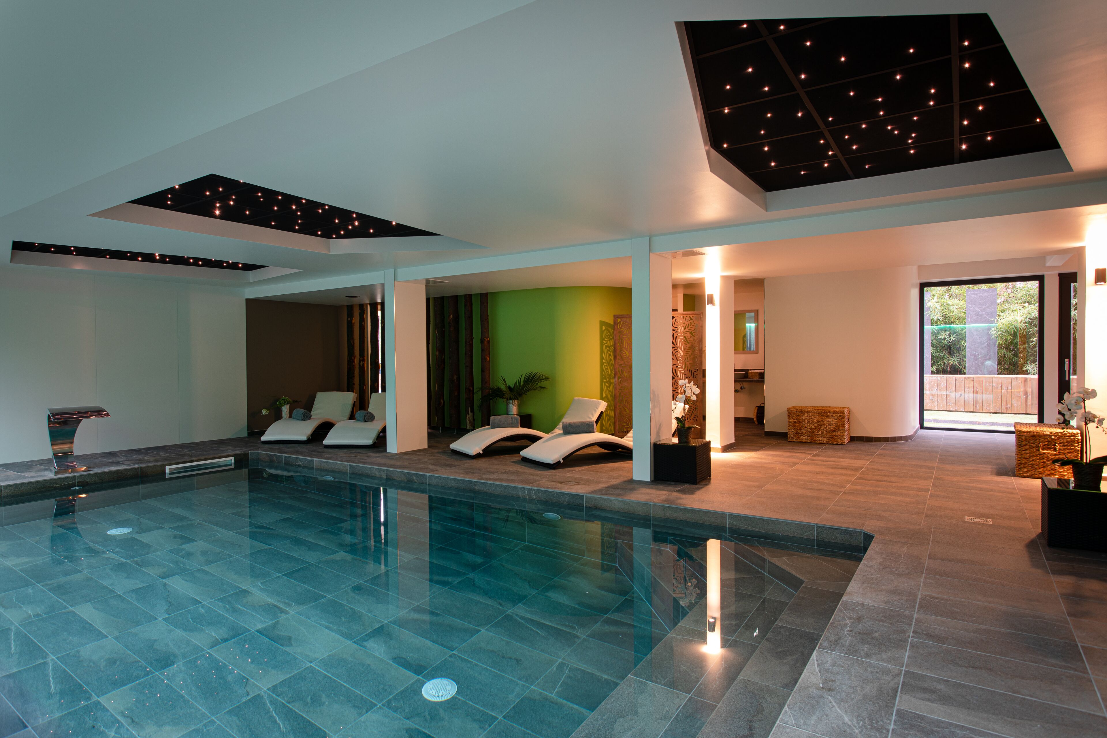 A modern indoor swimming pool with lounge chairs, a green accent wall, and recessed lighting.