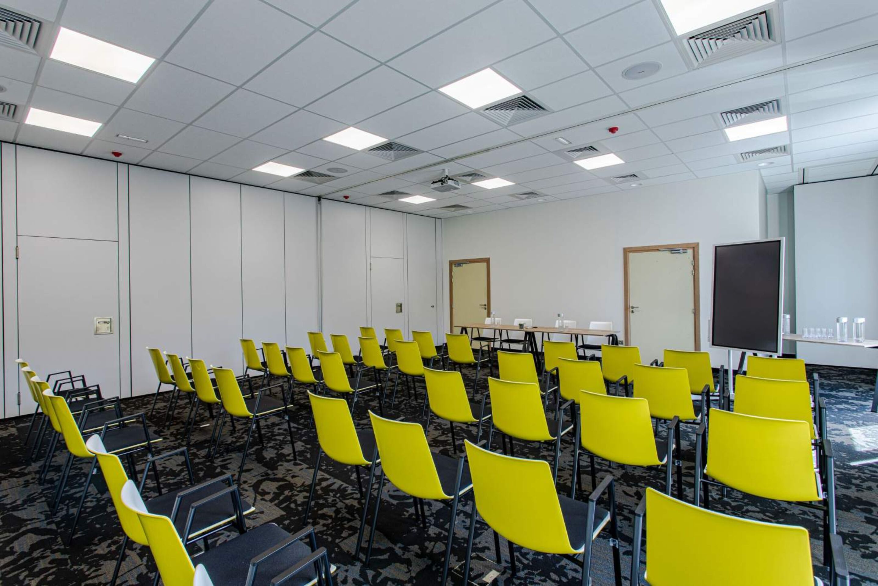 A conference room with rows of yellow chairs, a whiteboard, and a projector screen.