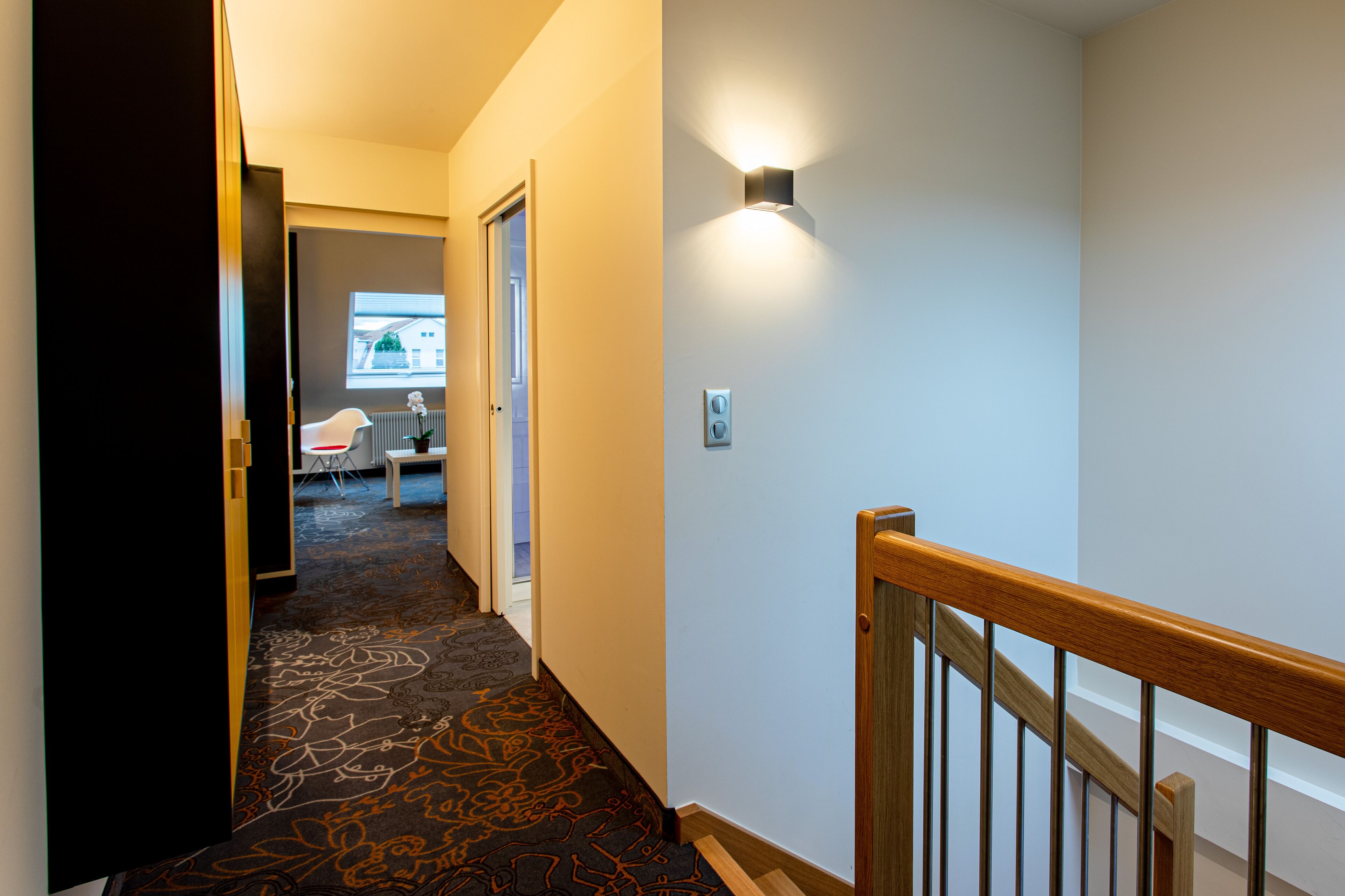 A hotel hallway with a carpeted floor, a wooden railing, and a light fixture on the wall.