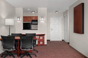 Una habitación de hotel con una mesa de comedor, sillas, una lámpara, una zona de cocina y una puerta.