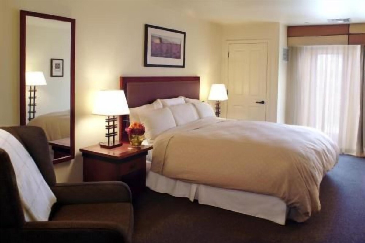 A hotel room with a bed, two bedside tables with lamps, a chair, a door, and a window with curtains.
