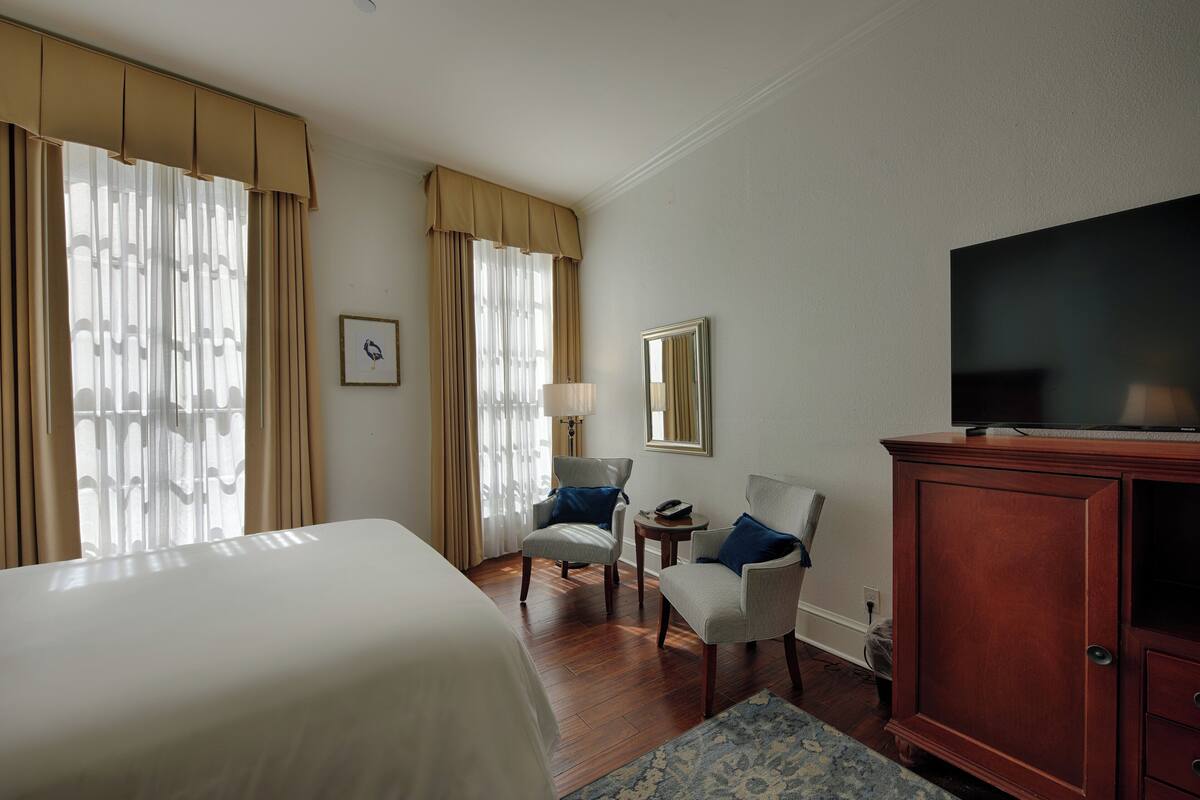 Habitación de hotel con una cama grande, dos sillones, un aparador de madera, un televisor y una mesita de noche con lámpara.