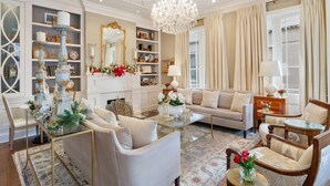 Ein geräumiges Wohnzimmer mit einem beigen Sofa, einem Glastisch und einer Kristallleuchter. Auf Regalen stehen Dekorationen und es gibt einen Kamin.