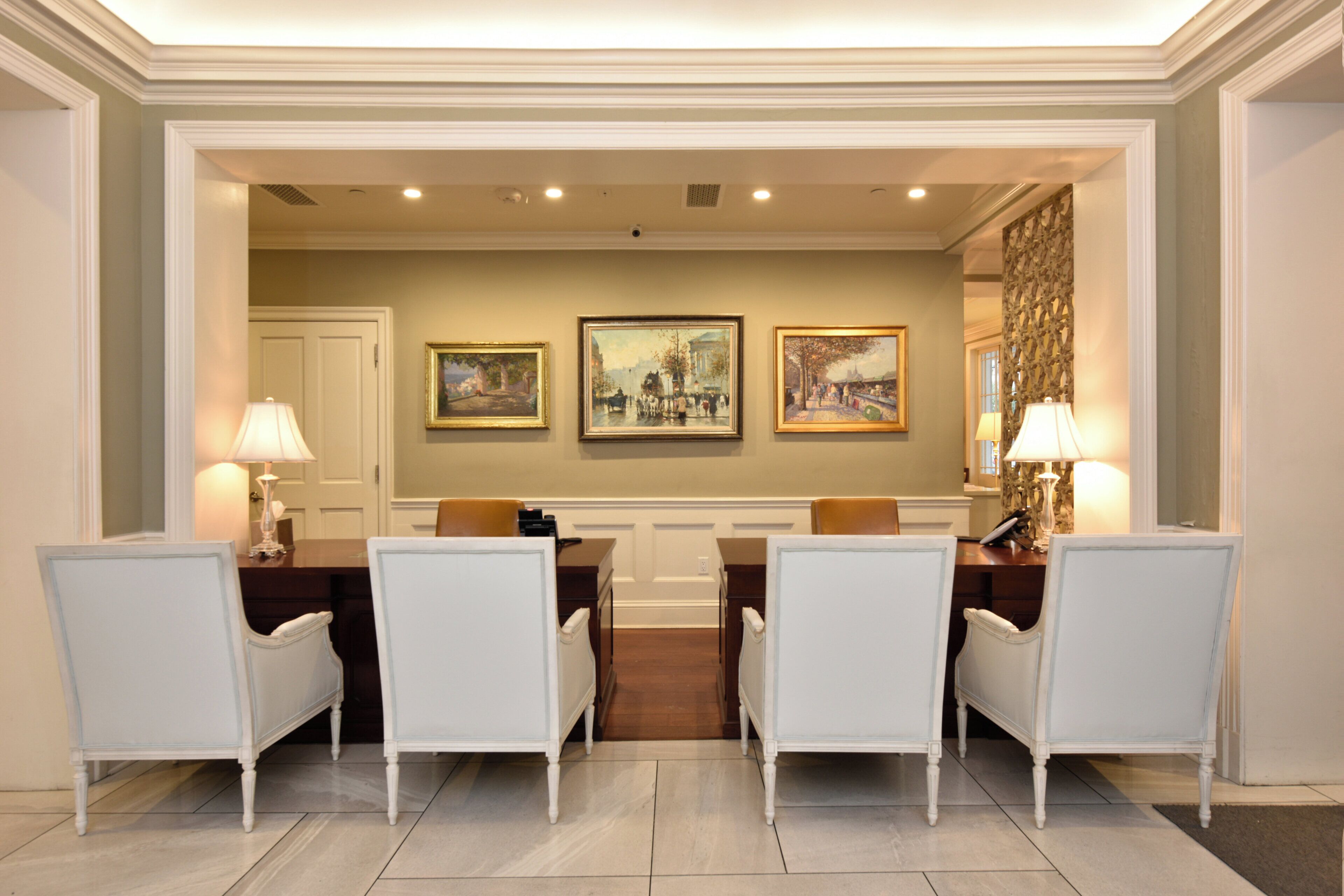 A formal dining room with a white table set for four, flanked by two white chairs, and three framed paintings on the wall.