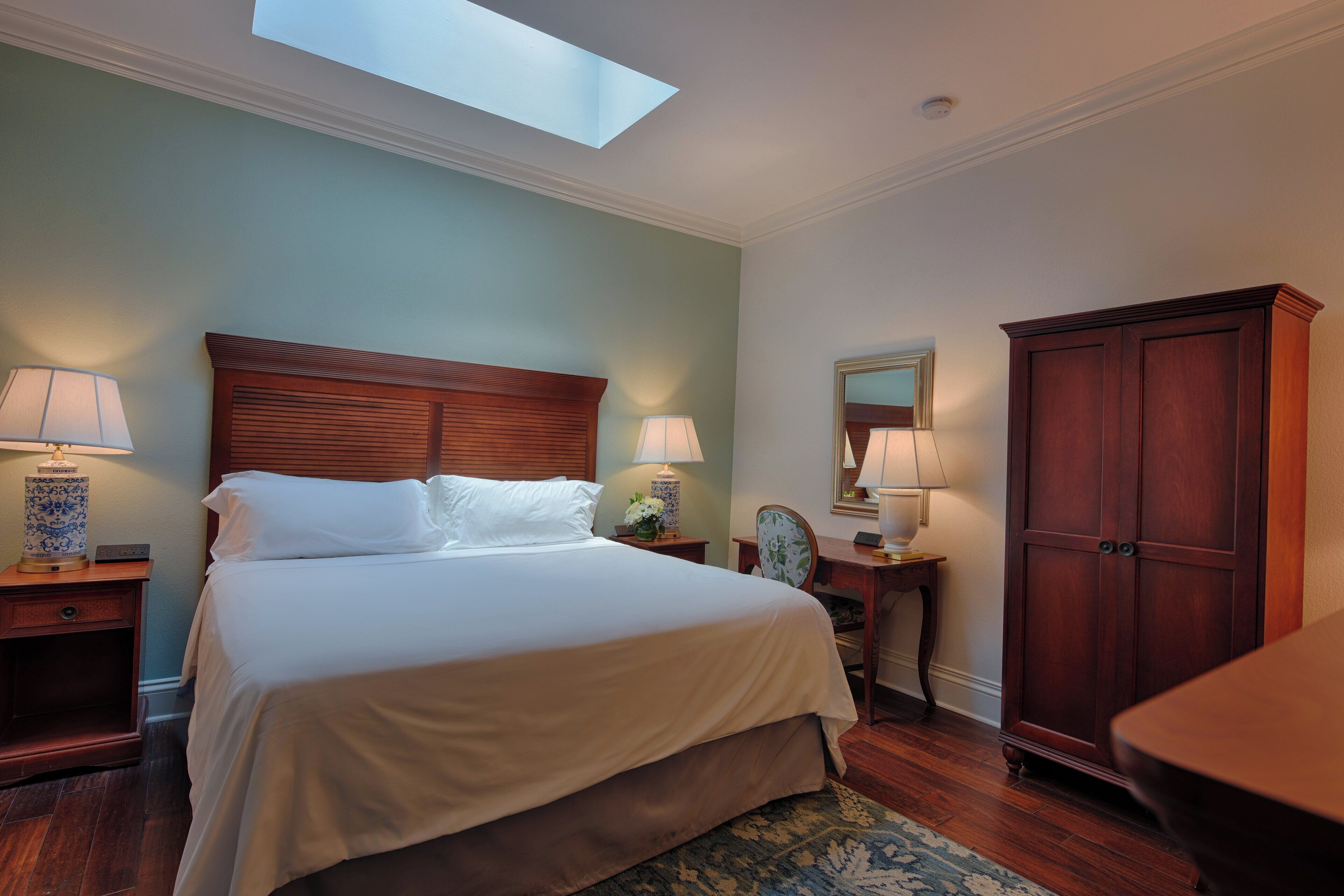 Un dormitorio ordenado con una cama grande, dos mesitas de noche con lámparas, un armario de madera y un tragaluz sobre la cama.