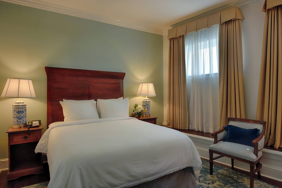 Habitación de hotel con una cama grande, dos mesitas de noche con lámparas, una silla y una ventana con cortinas.