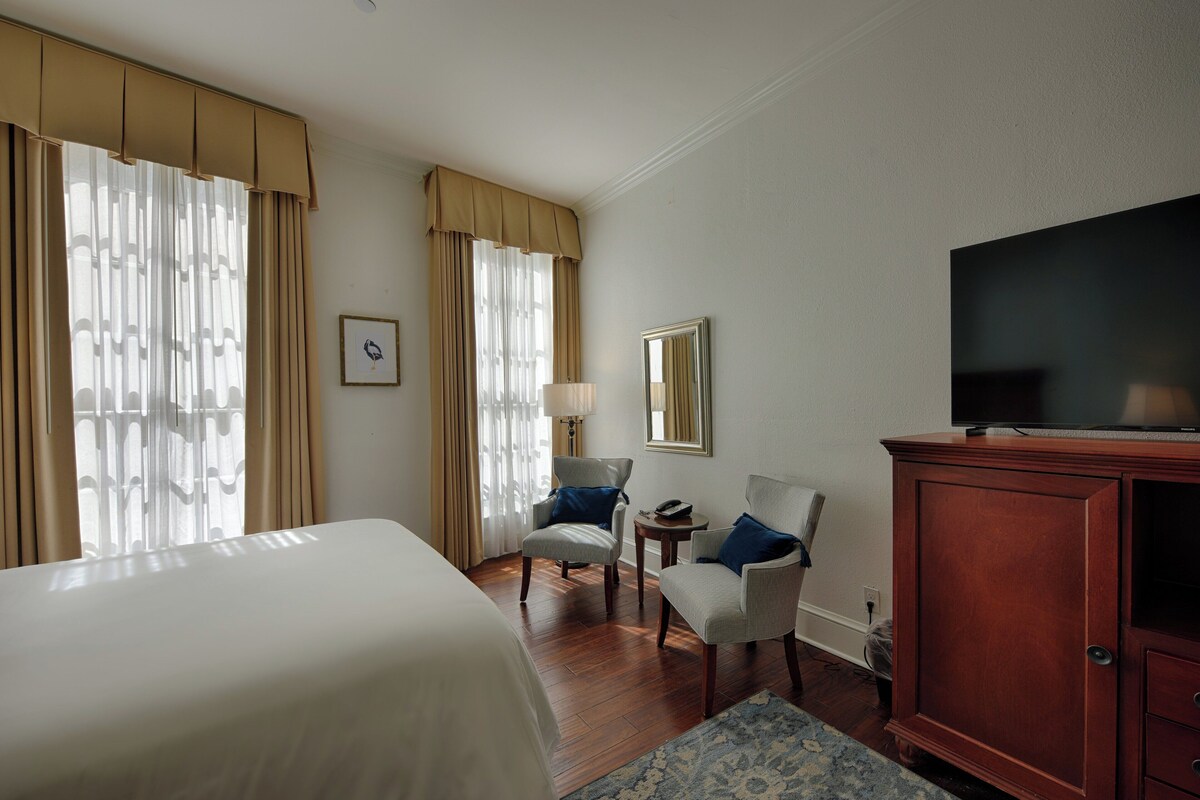Habitación de hotel con una cama grande, dos sillones, un aparador de madera, un televisor y una mesita de noche con lámpara.