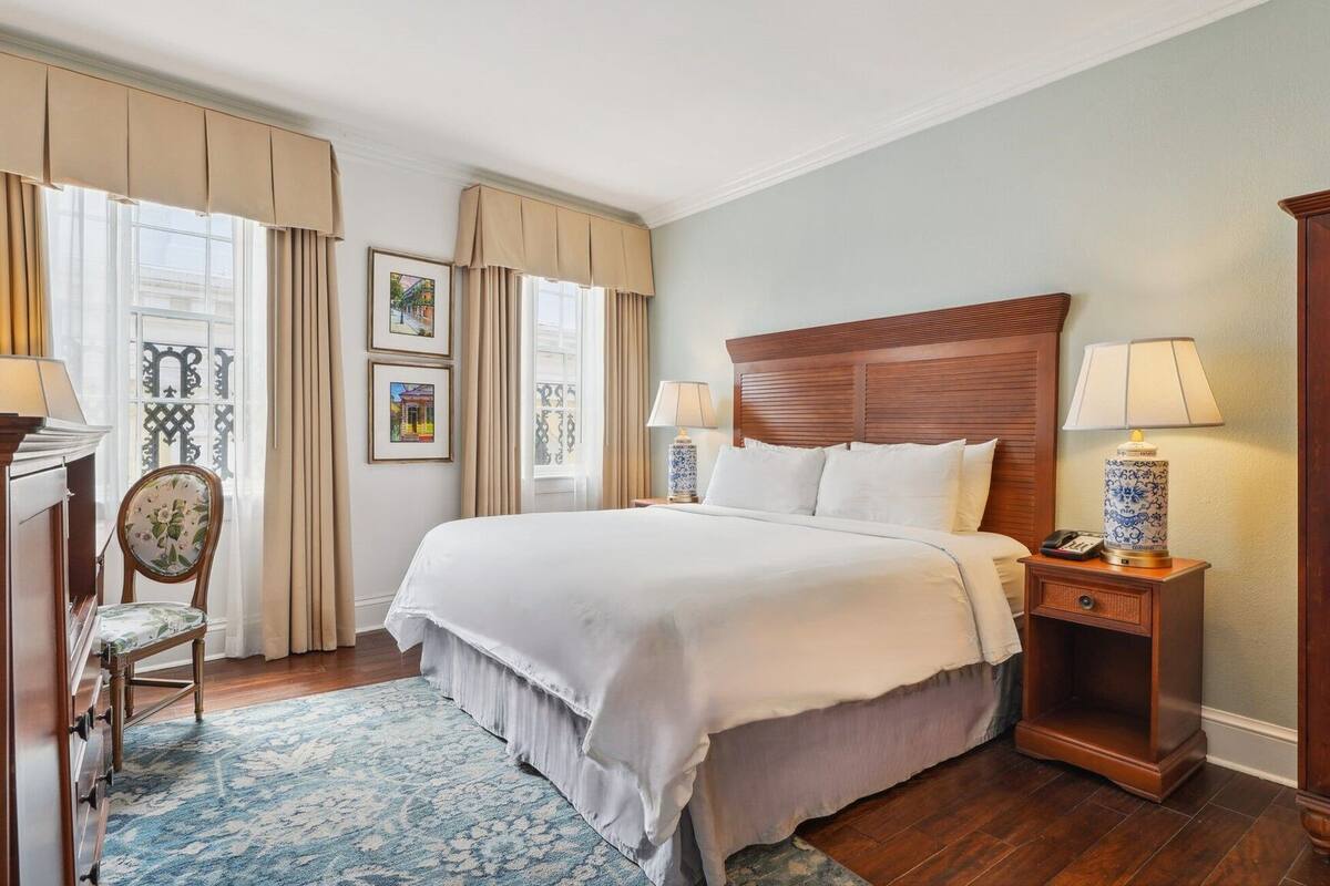 Una habitación de hotel ordenada con una cama grande, una mesita de noche de madera con una lámpara, una silla con un cojín estampado y una ventana con cortinas.
