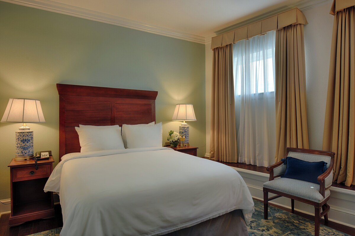 Habitación de hotel con una cama grande, dos mesitas de noche con lámparas, una silla y una ventana con cortinas.