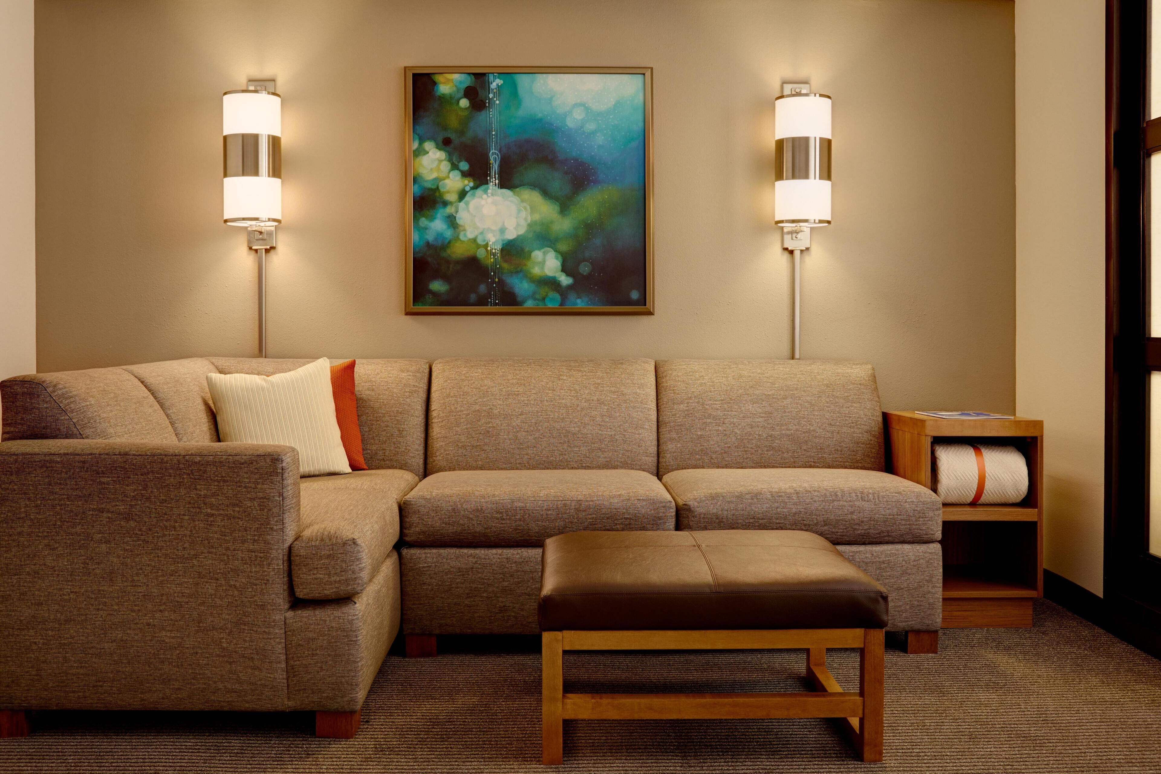 A modern living room with a sectional sofa, a wooden bench, two wall-mounted lamps, and a framed abstract painting.