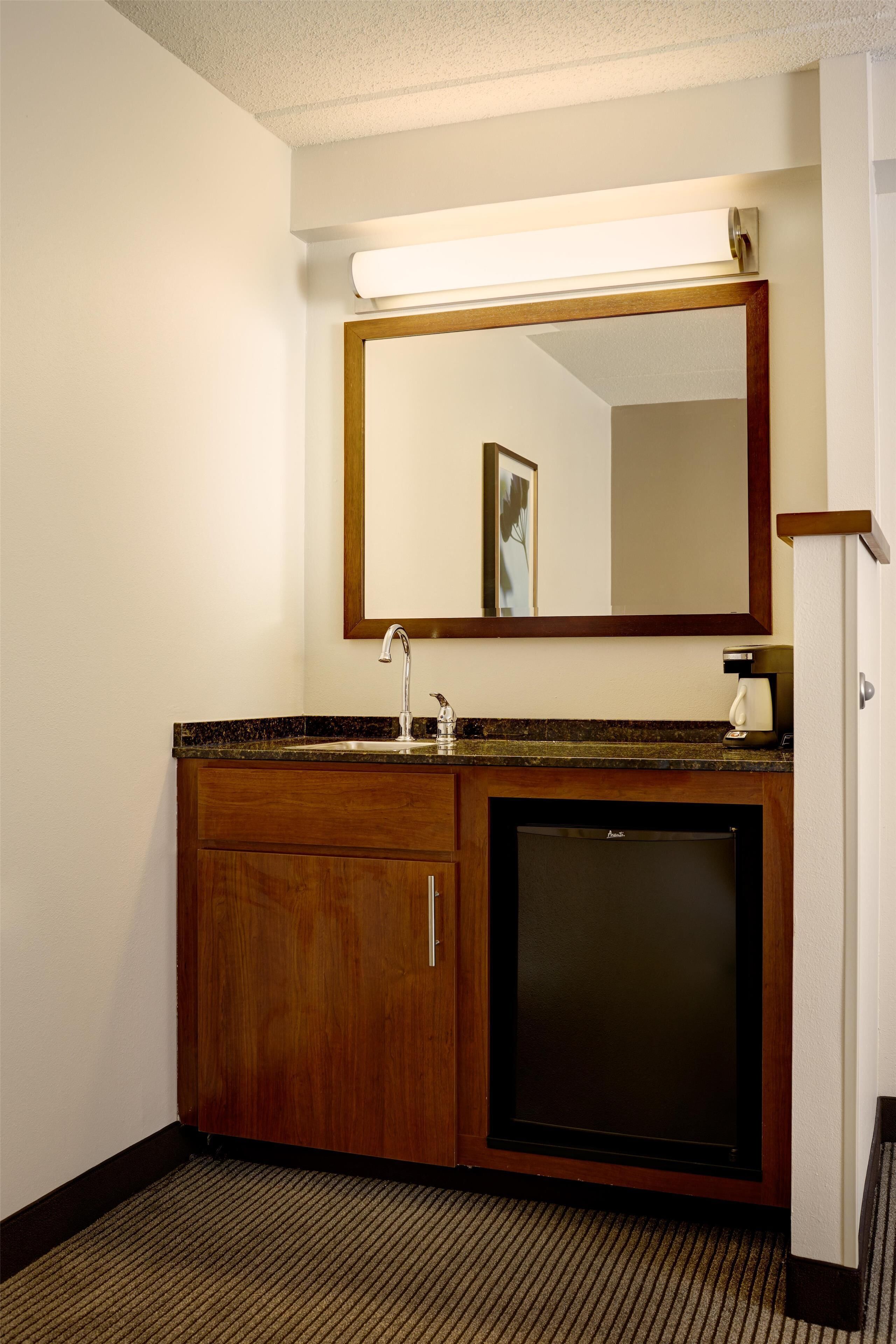 A hotel bathroom with a wooden vanity, a sink, a mirror, and a refrigerator.