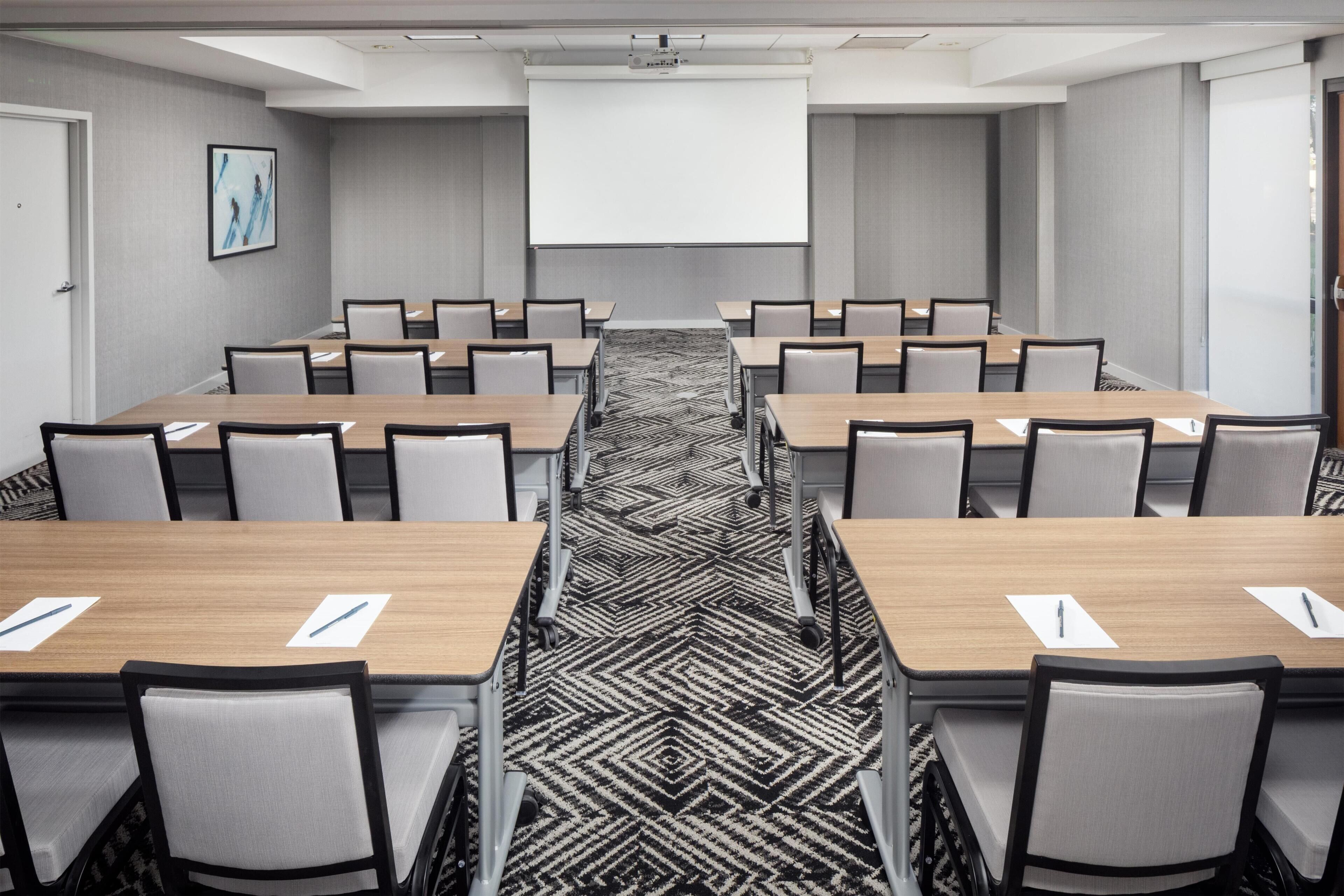 A conference room with a long table, chairs, a projector screen, and a patterned carpet.