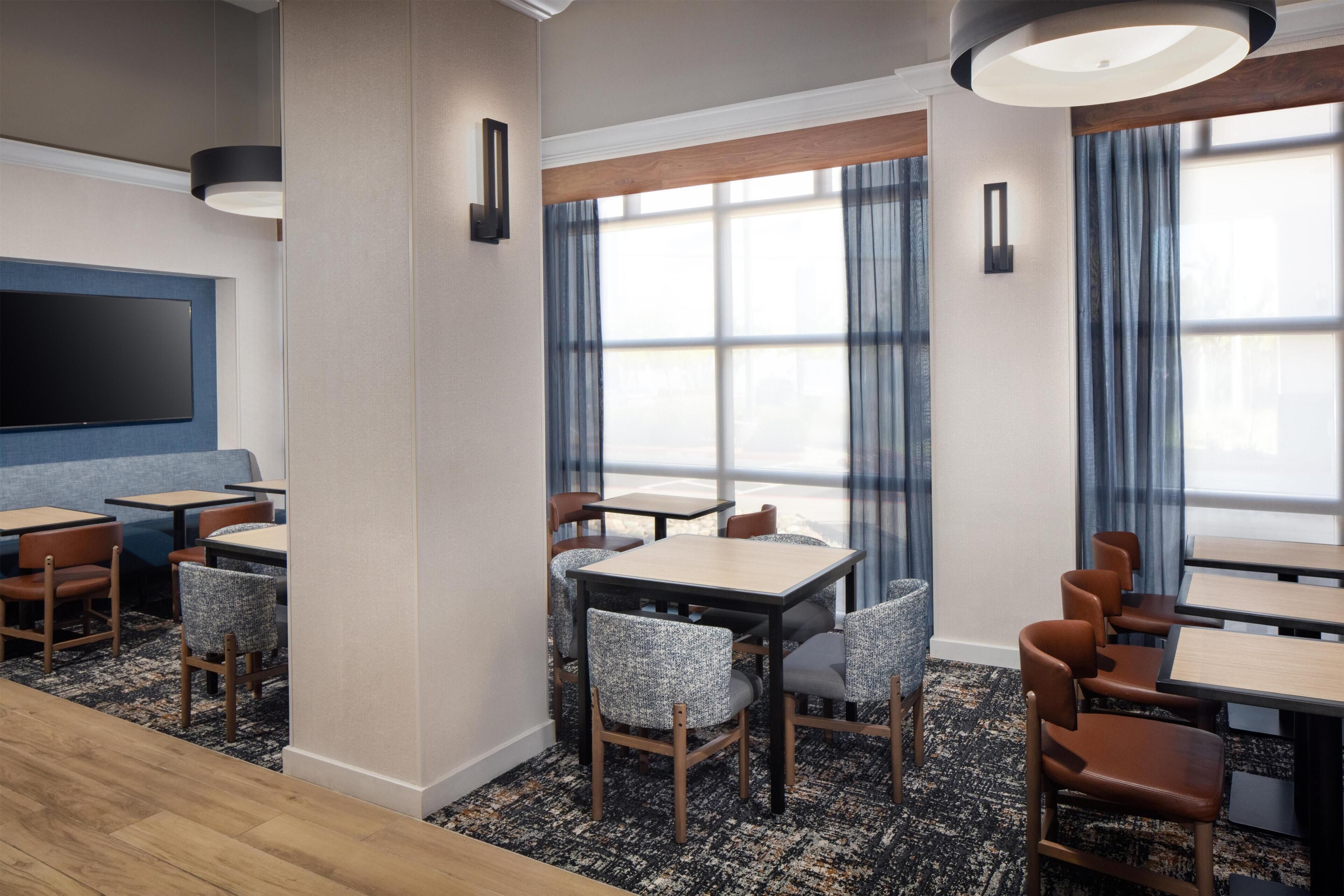 A hotel room with a flat-screen TV, multiple dining tables, chairs, and a large window with curtains.