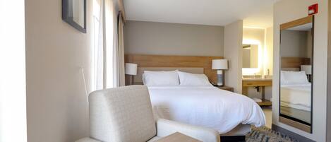 A hotel room with a bed, a seating area, a bedside table, a mirror, and a window with curtains.