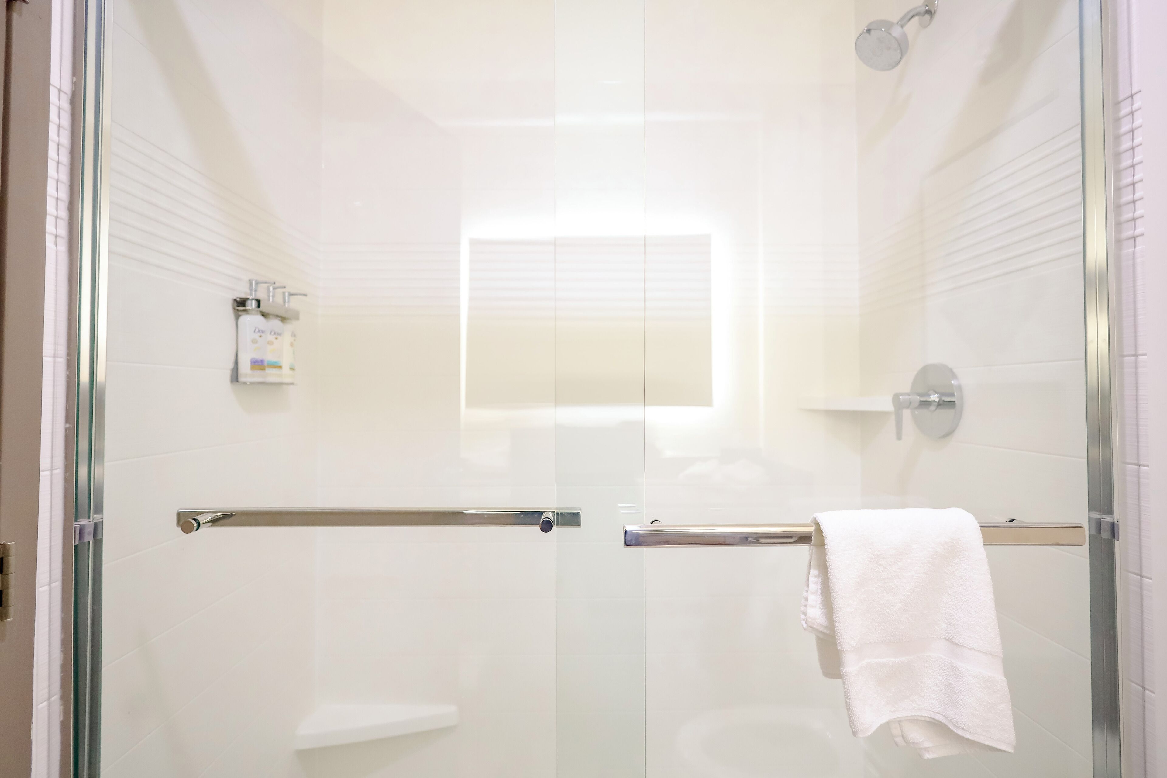A modern bathroom with a glass shower enclosure, a white towel hanging on a towel bar, and a soap dispenser on a shelf.