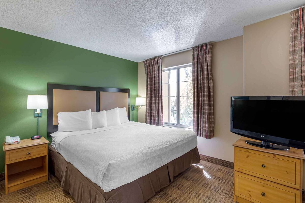 Une chambre d’hôtel avec un grand lit, un téléviseur posé sur un meuble en bois, une table de chevet avec une lampe et une fenêtre avec des rideaux.