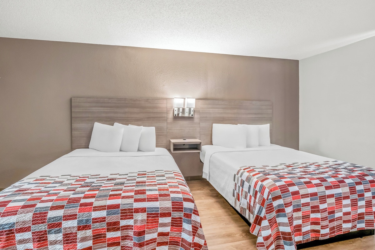 A hotel room with two beds, each with a different patterned bedspread. There is a nightstand with a lamp between the beds, and a wall-mounted light fixture above it.
