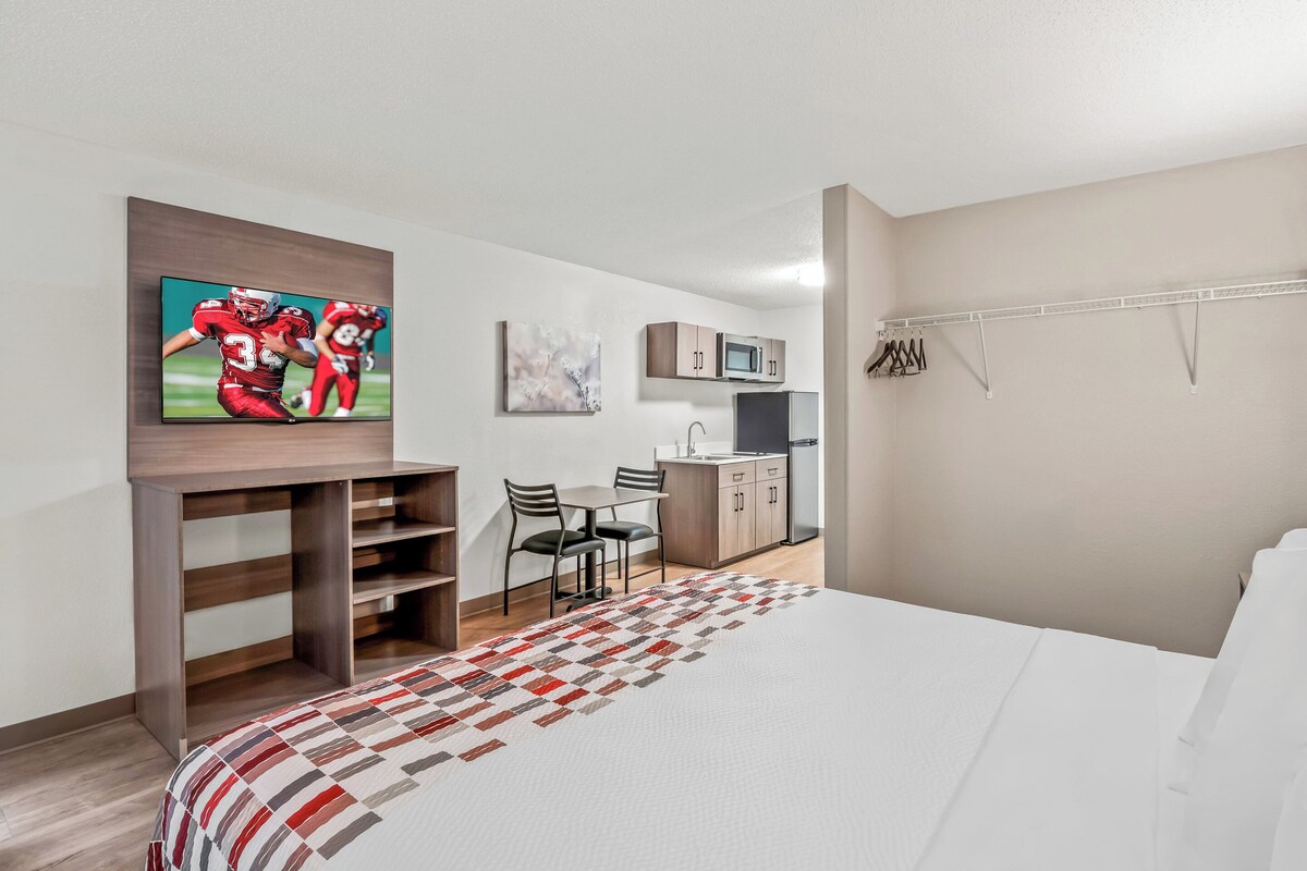 A hotel room with a bed, a TV mounted on a wooden shelf, a kitchenette, and a dining area with chairs.