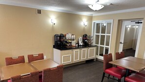 A dining area with tables and chairs, a coffee station with various coffee makers, and a door leading to another room. - Red Roof Inn Southfield (Southfield)