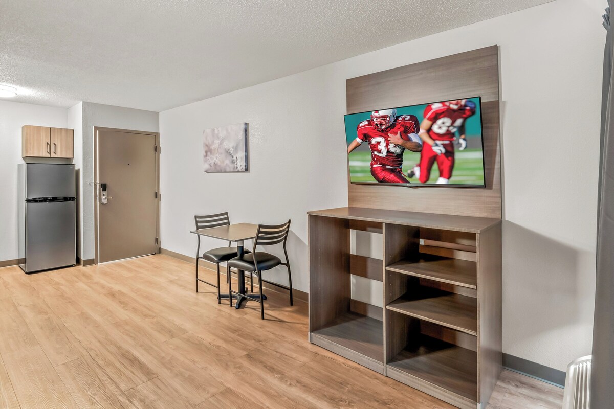 A modern living room with a flat-screen TV, wooden flooring, a dining table with chairs, and a built-in shelving unit.