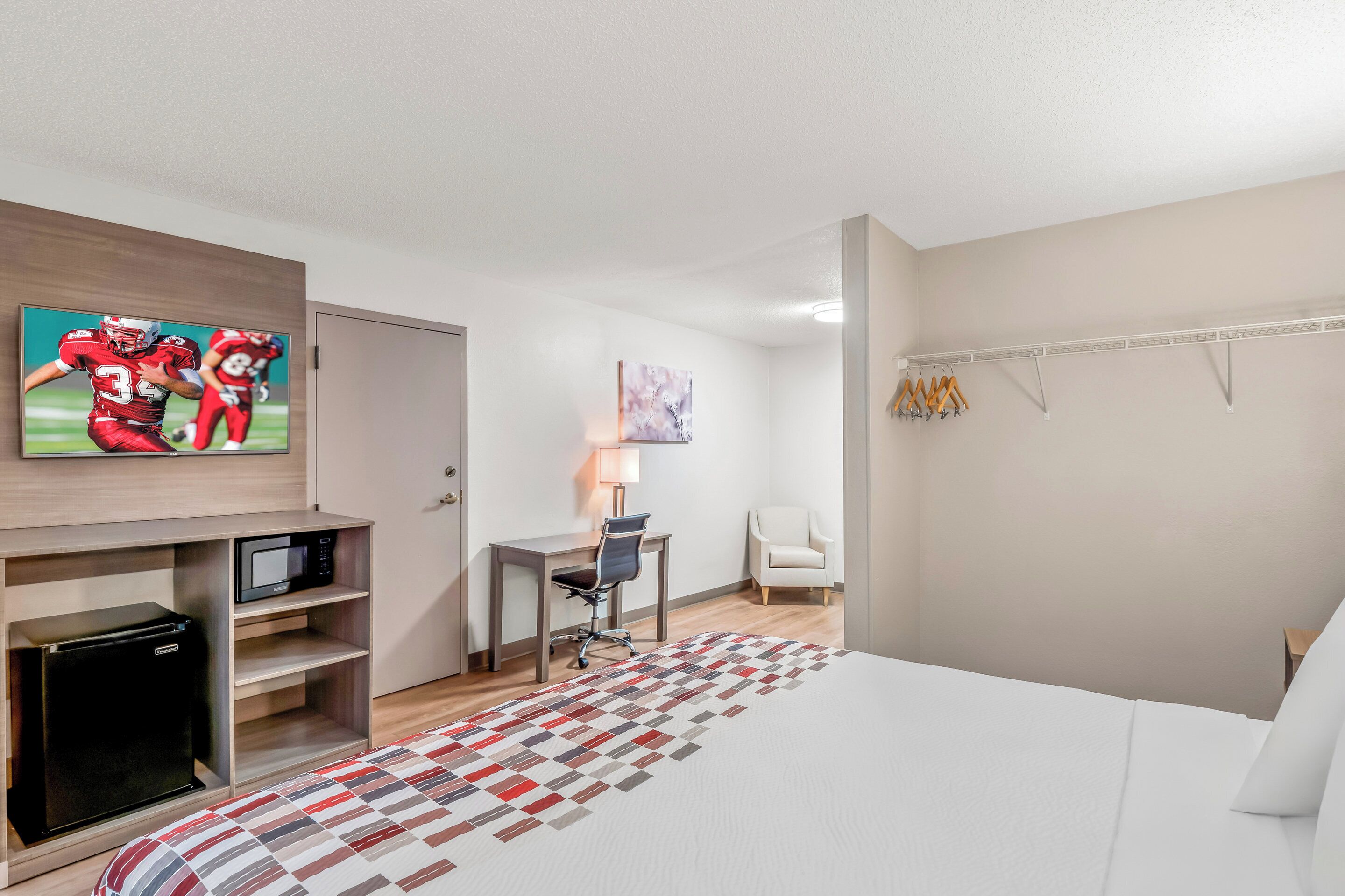 Habitación de hotel con cama, televisión montada en la pared, escritorio con silla, refrigerador pequeño y perchero.