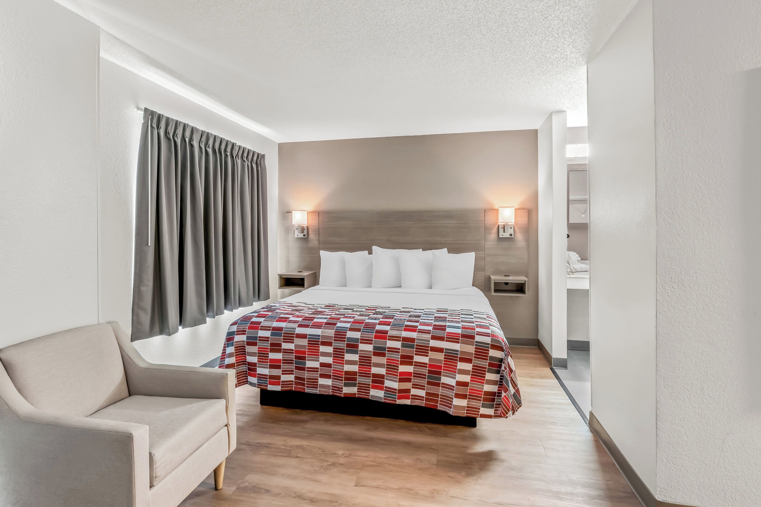 A hotel room with a bed, a chair, a nightstand, and a wall-mounted lamp.